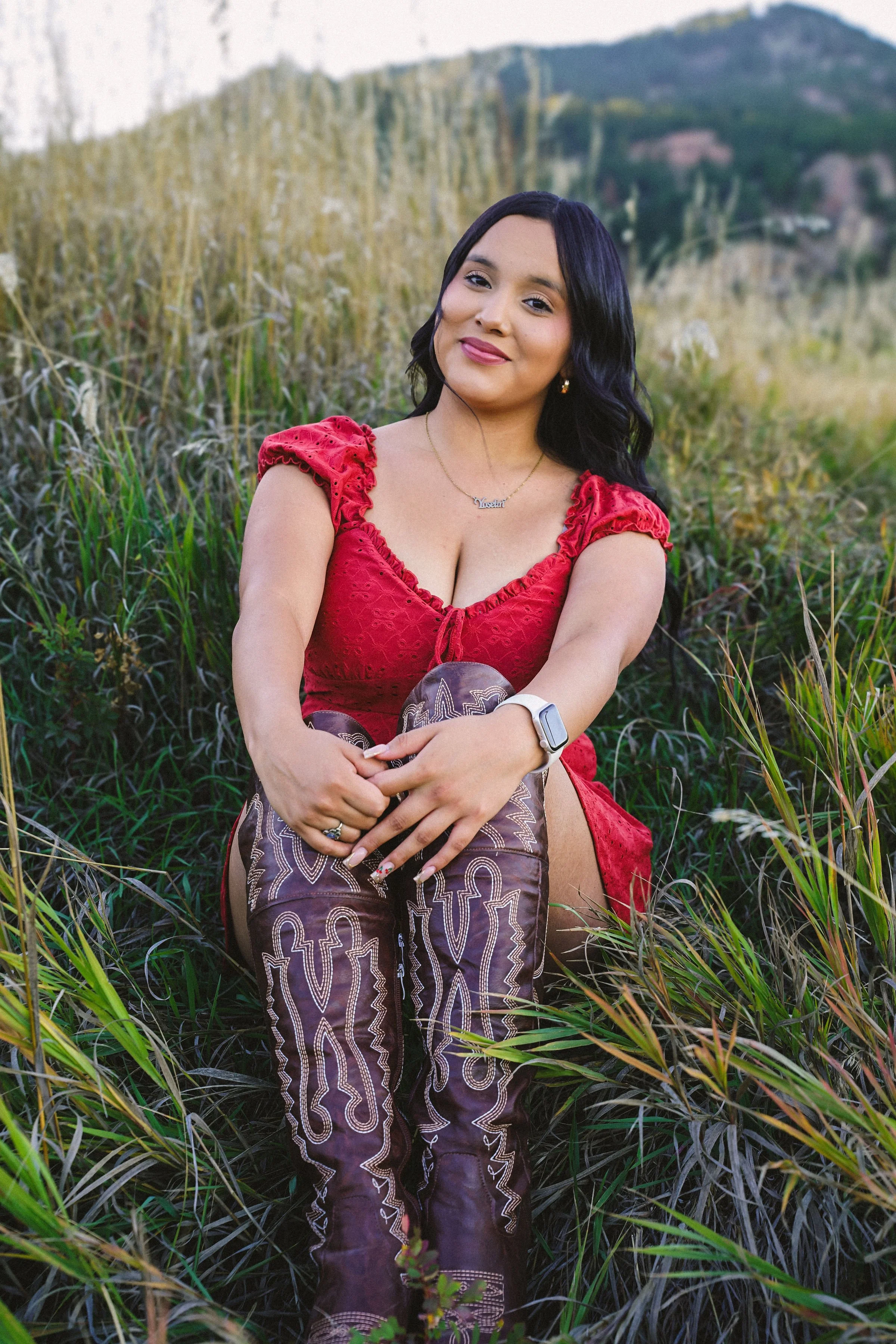 Woman in a red dress sitting in a field of tall grass with hills in the background.