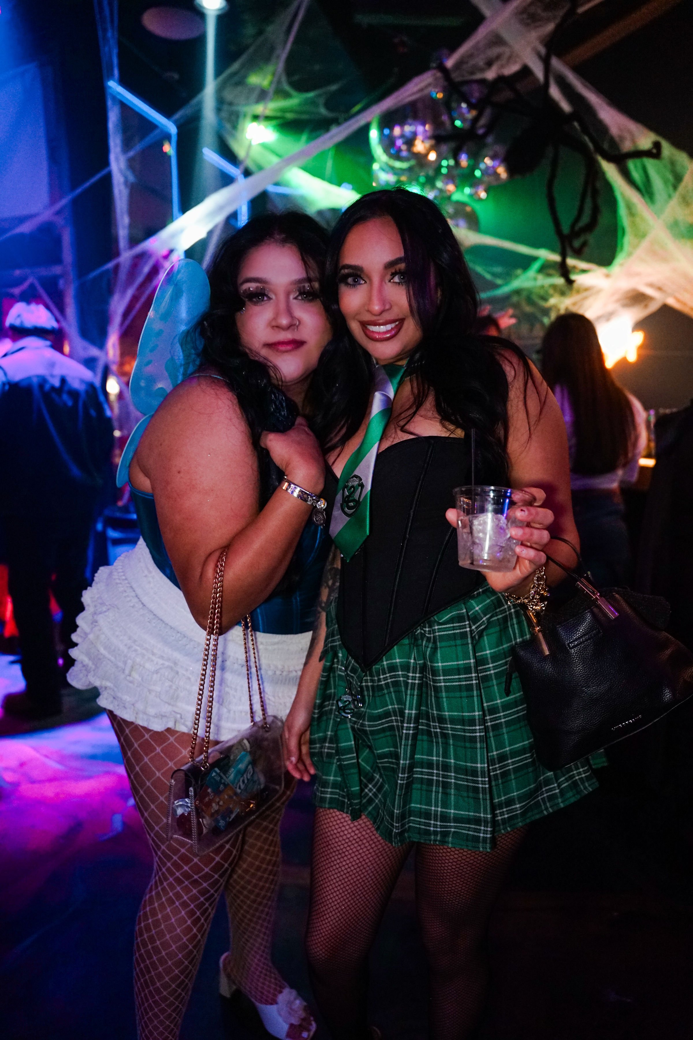 Two women at a party dressed in costumes with colorful lighting, Halloween decorations, and cobwebs in the background.