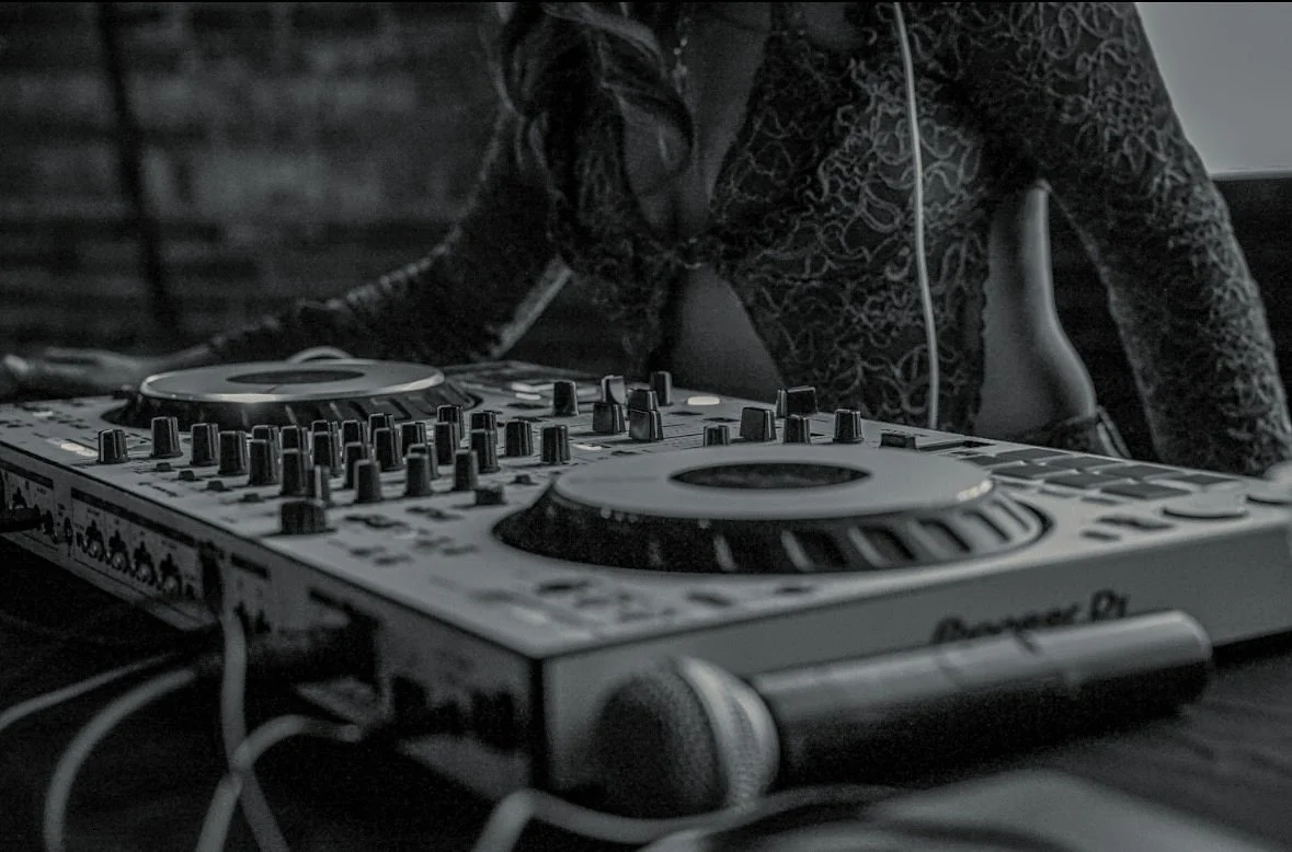 Close-up of a DJ wearing headphones using a professional DJ controller with multiple knobs and jog wheels in a dimly lit setting.