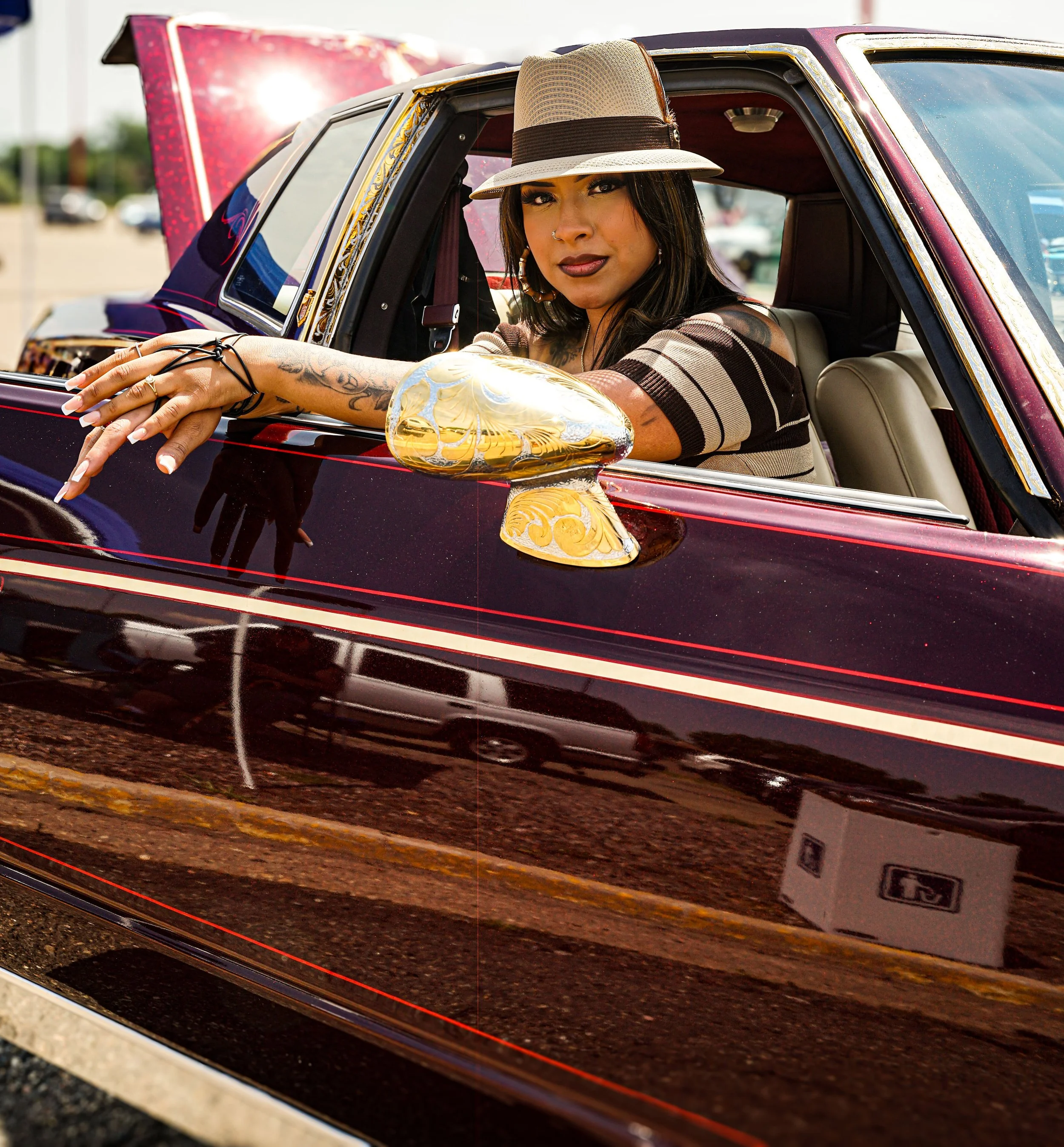 Woman with tattoos and wearing a hat sitting in a vintage car with a shiny, decorative side mirror, in an outdoor parking lot.