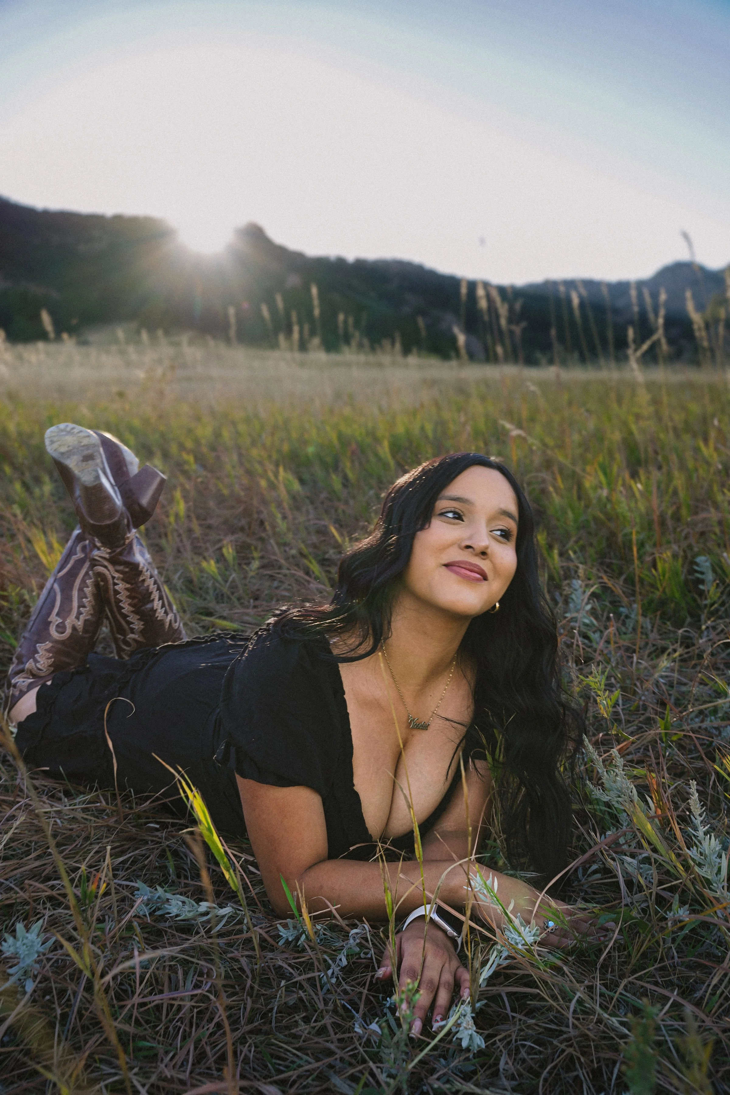 A woman with long black hair lying on her stomach in a grassy field during sunset, smiling slightly, wearing a black top, patterned boots, jewelry, and a smartwatch, with mountains in the background.