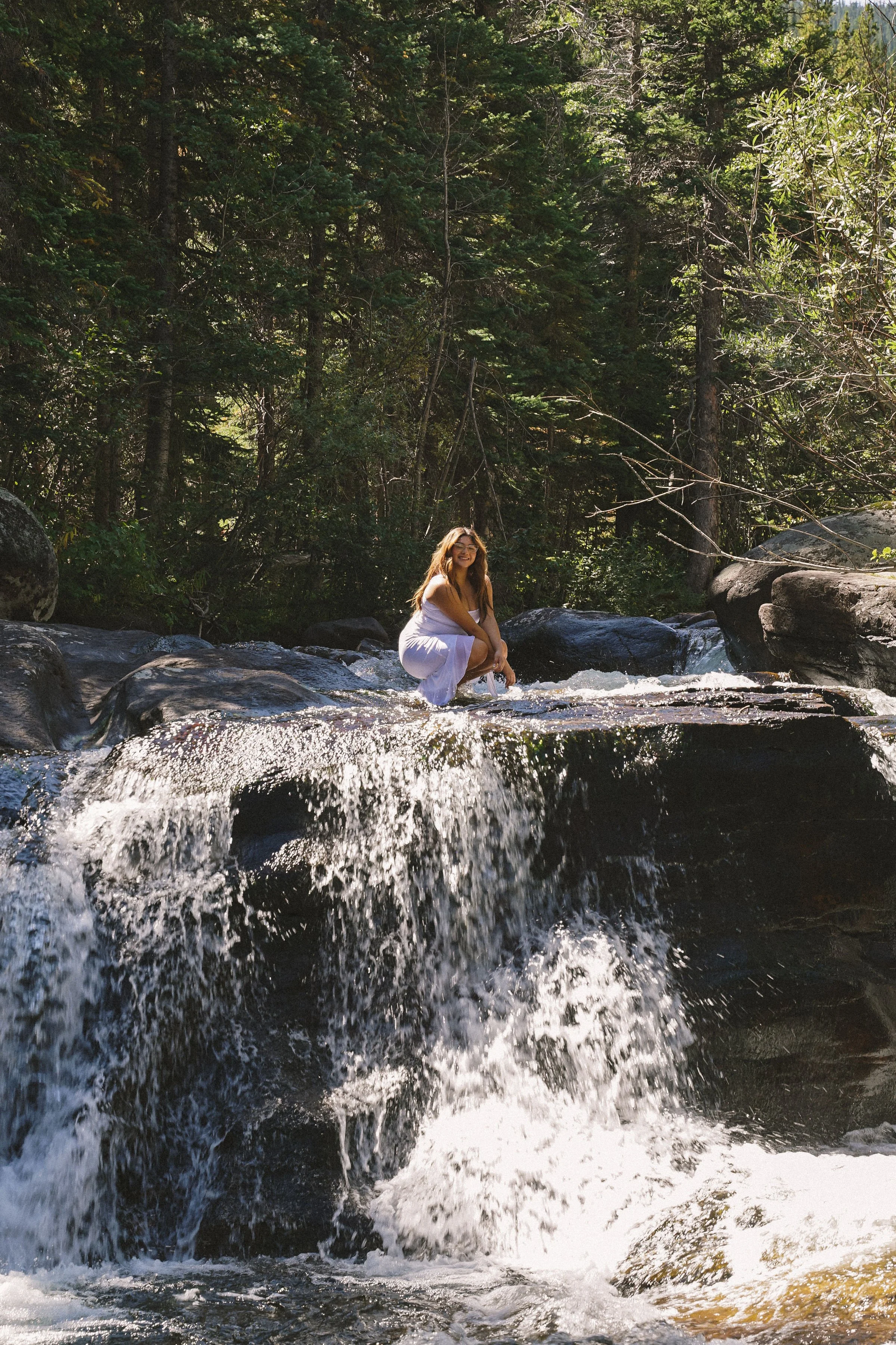 A woman in a white dress squatting on rocks in a stream with small waterfall surrounded by green trees.