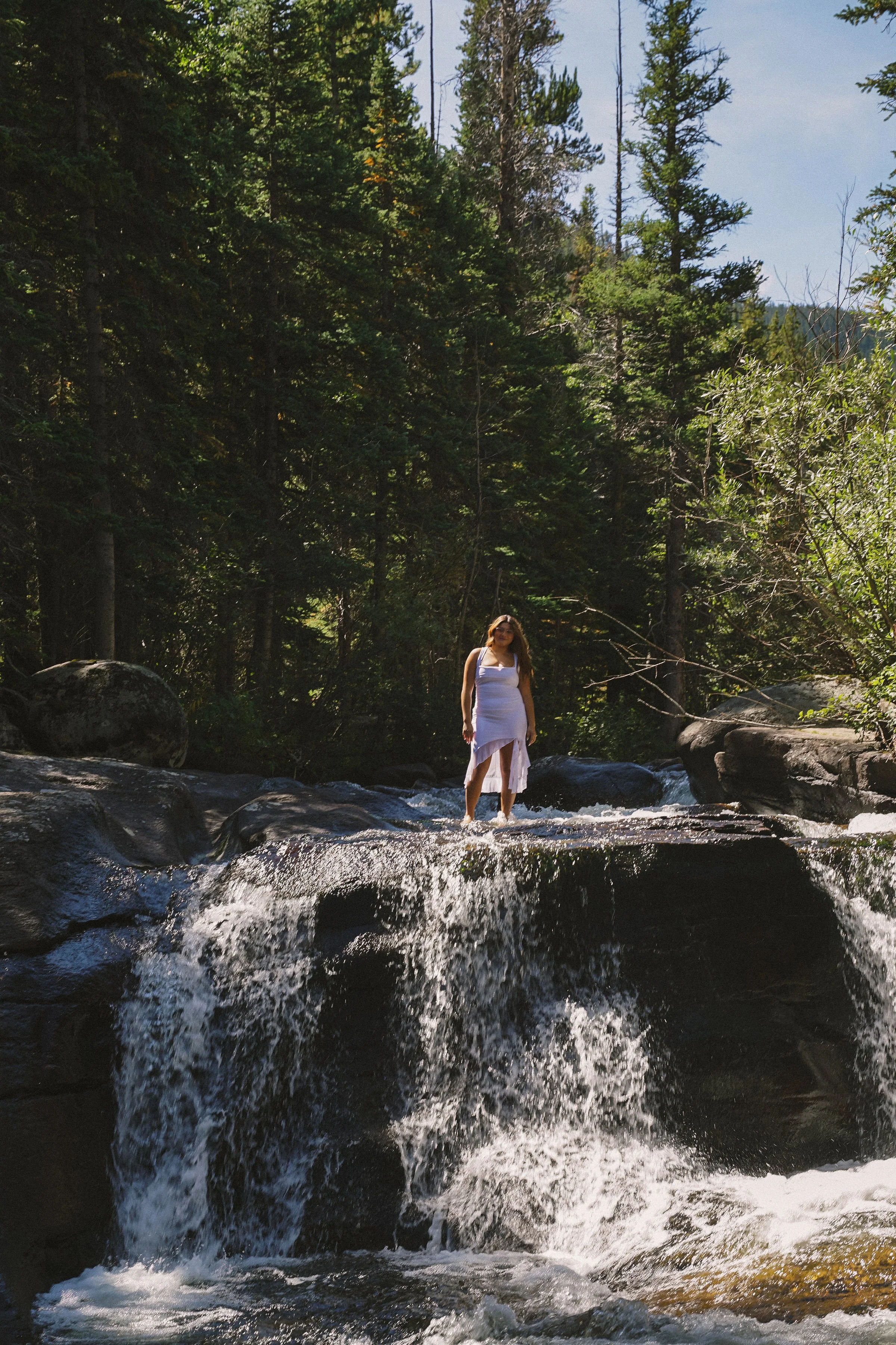 A woman in a white dress standing on rocks in a flowing river surrounded by tall green trees in a forested area.