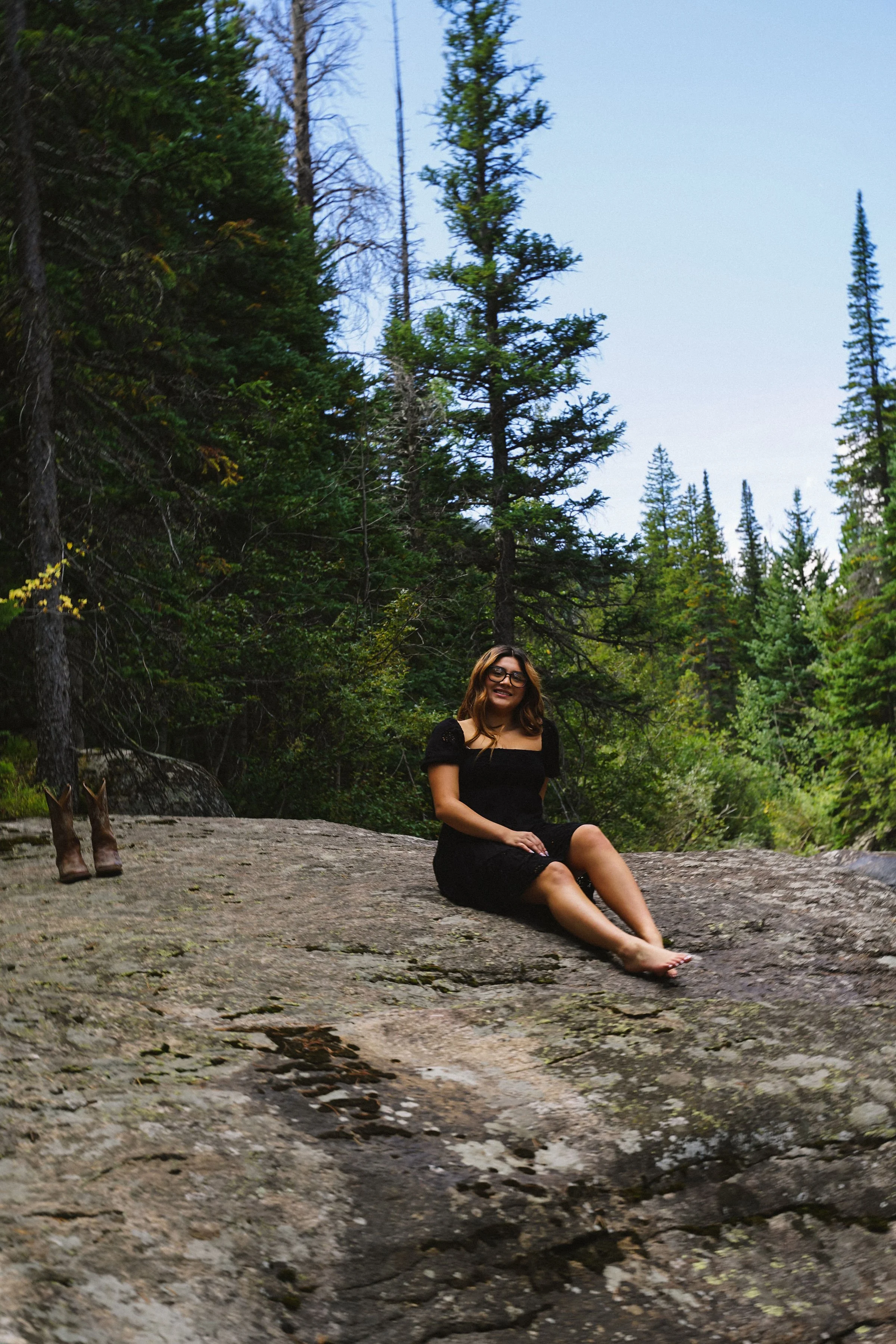 A woman with glasses and a black dress sits on a large rock in a forest, smiling, with trees in the background.