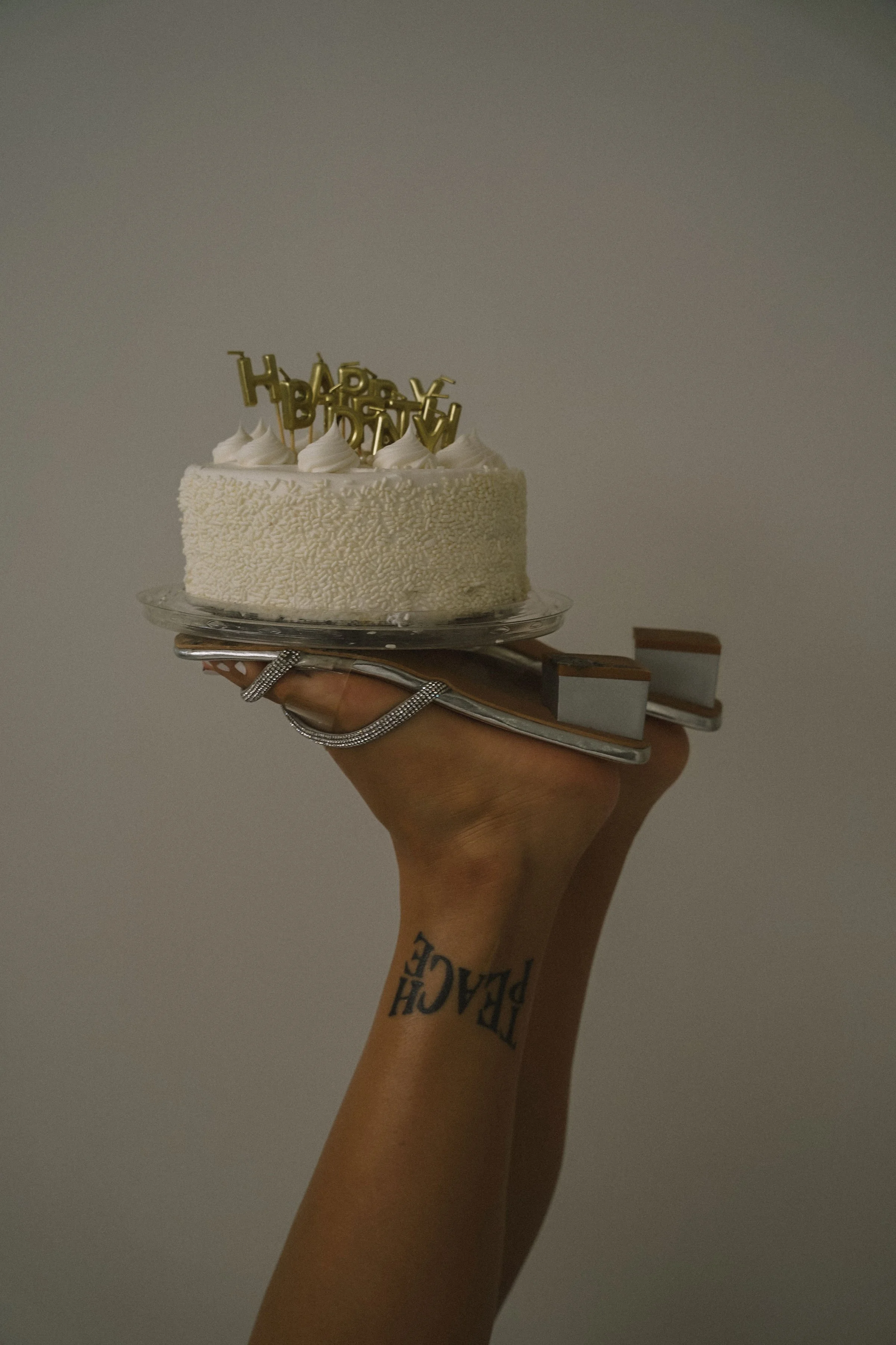 A hand with a tattoo on the wrist is holding a silver tray with a white frosted birthday cake topped with white icing and gold 'Happy Birthday' candles against a plain background.