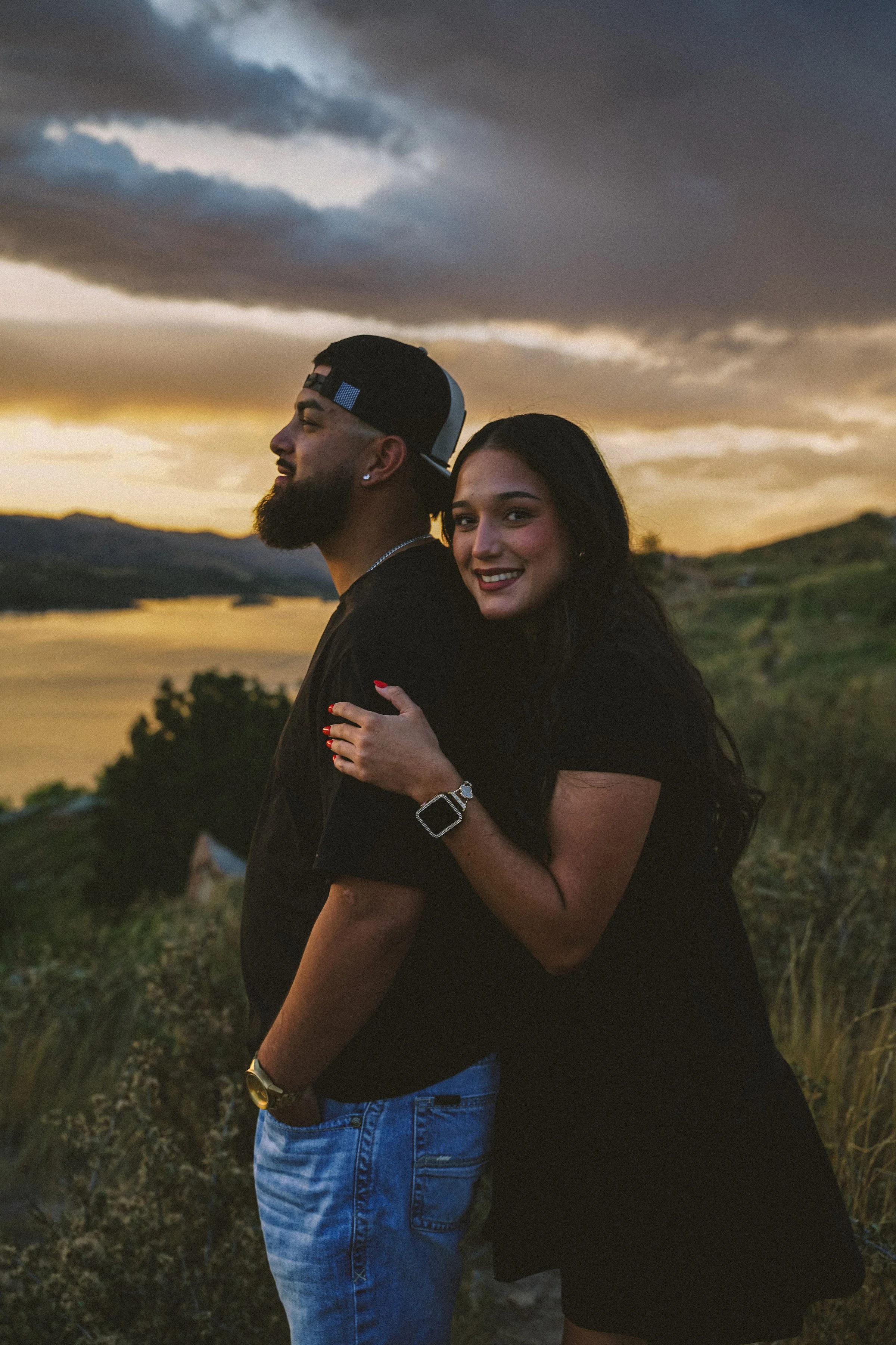 A smiling woman hugging a bearded man from behind during sunset next to a body of water and hilly landscape.