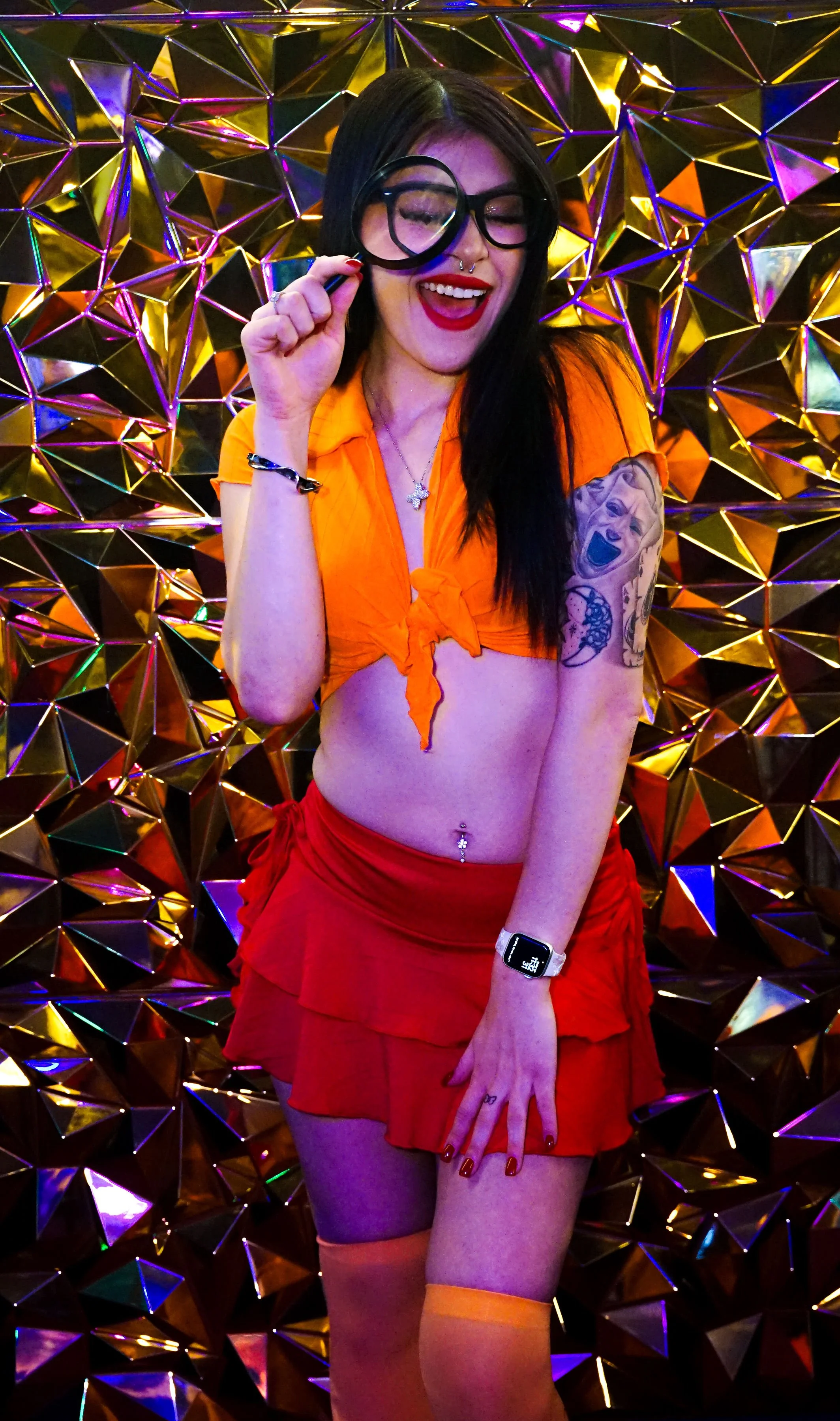 Woman with sunglasses holding a magnifying glass in front of her eye, wearing a bright orange tied-up top and a red ruffled skirt, in front of a metallic geometric background.