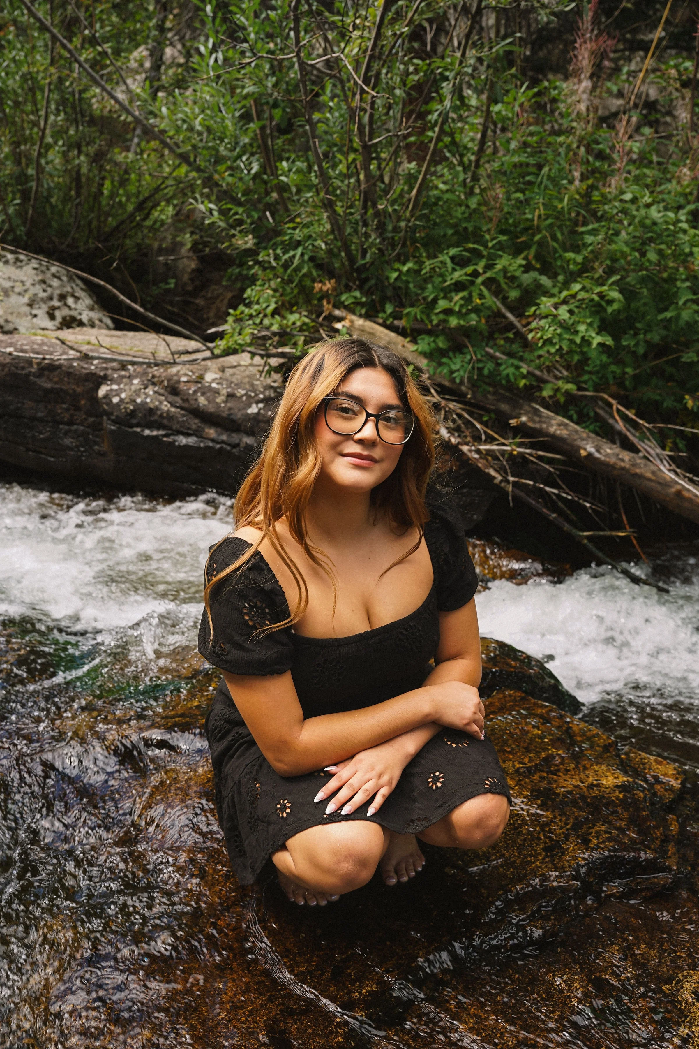 A woman with long wavy hair, glasses, and a black dress crouching on a rock in a flowing creek with green foliage in the background.