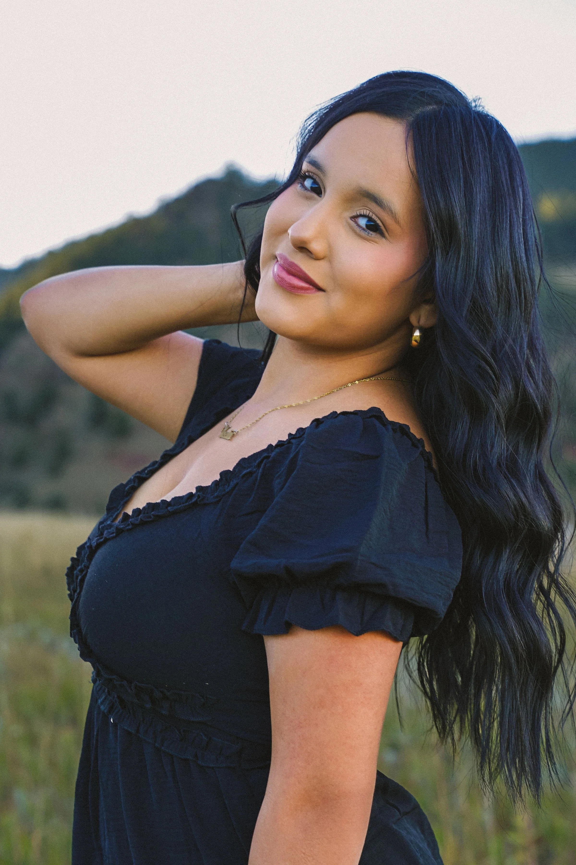 A young woman with long wavy black hair, wearing a black dress with puffed sleeves, posing outdoors in a field with trees and hills in the background, during sunset.