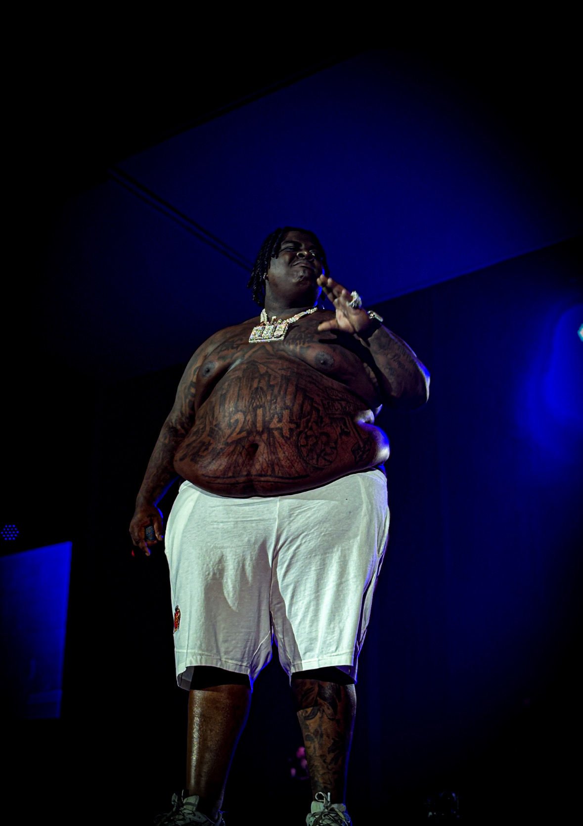 A shirtless man with tattoos standing on a stage with blue lighting, wearing white shorts, sneakers, and jewelry.