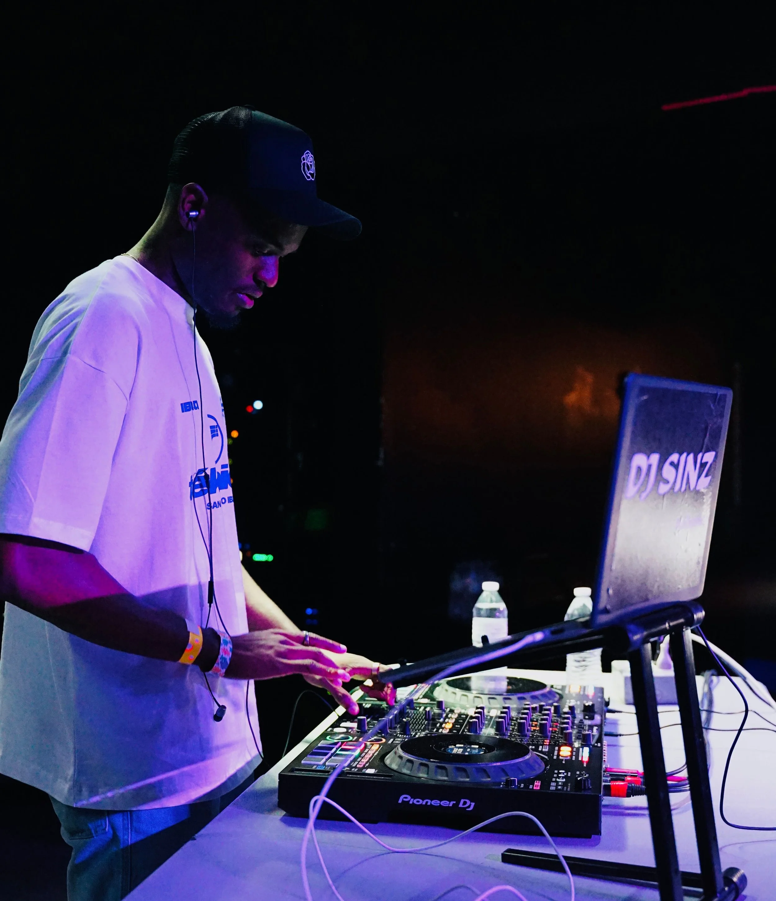 A DJ wearing a baseball cap, white T-shirt, and earphones is mixing music on a Pioneer DJ controller at a dark venue, with bottles of water on the table.