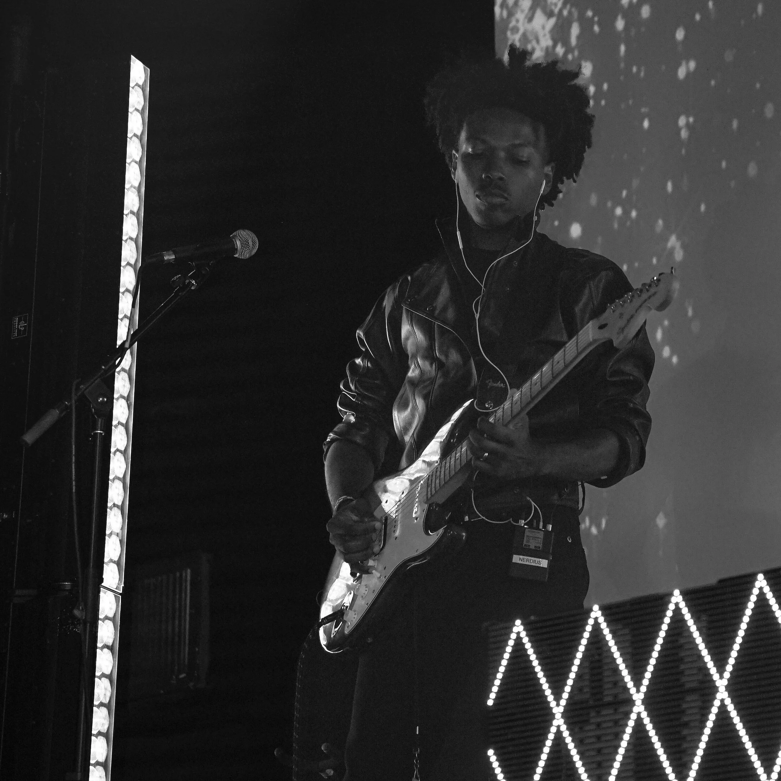 A person playing an electric guitar on stage, wearing a leather jacket and headphones, with a microphone stand nearby and a digital display with a diamond pattern in the background.