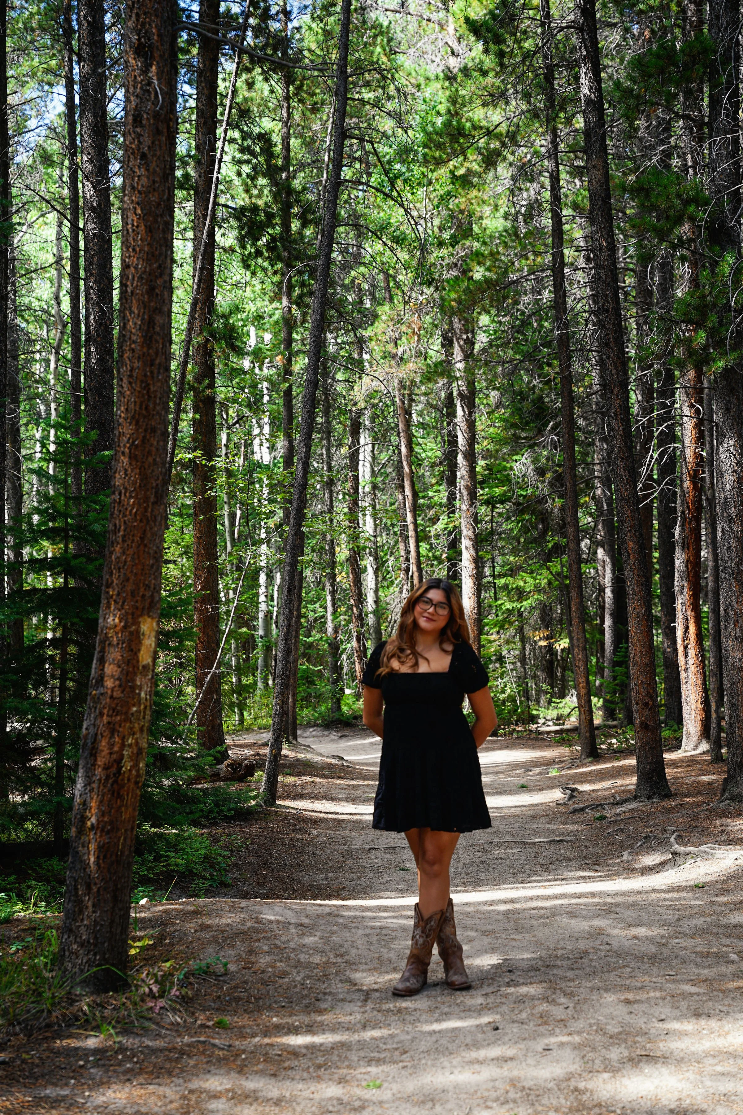 A woman in a black dress and cowboy boots standing on a dirt forest trail surrounded by tall pine trees.