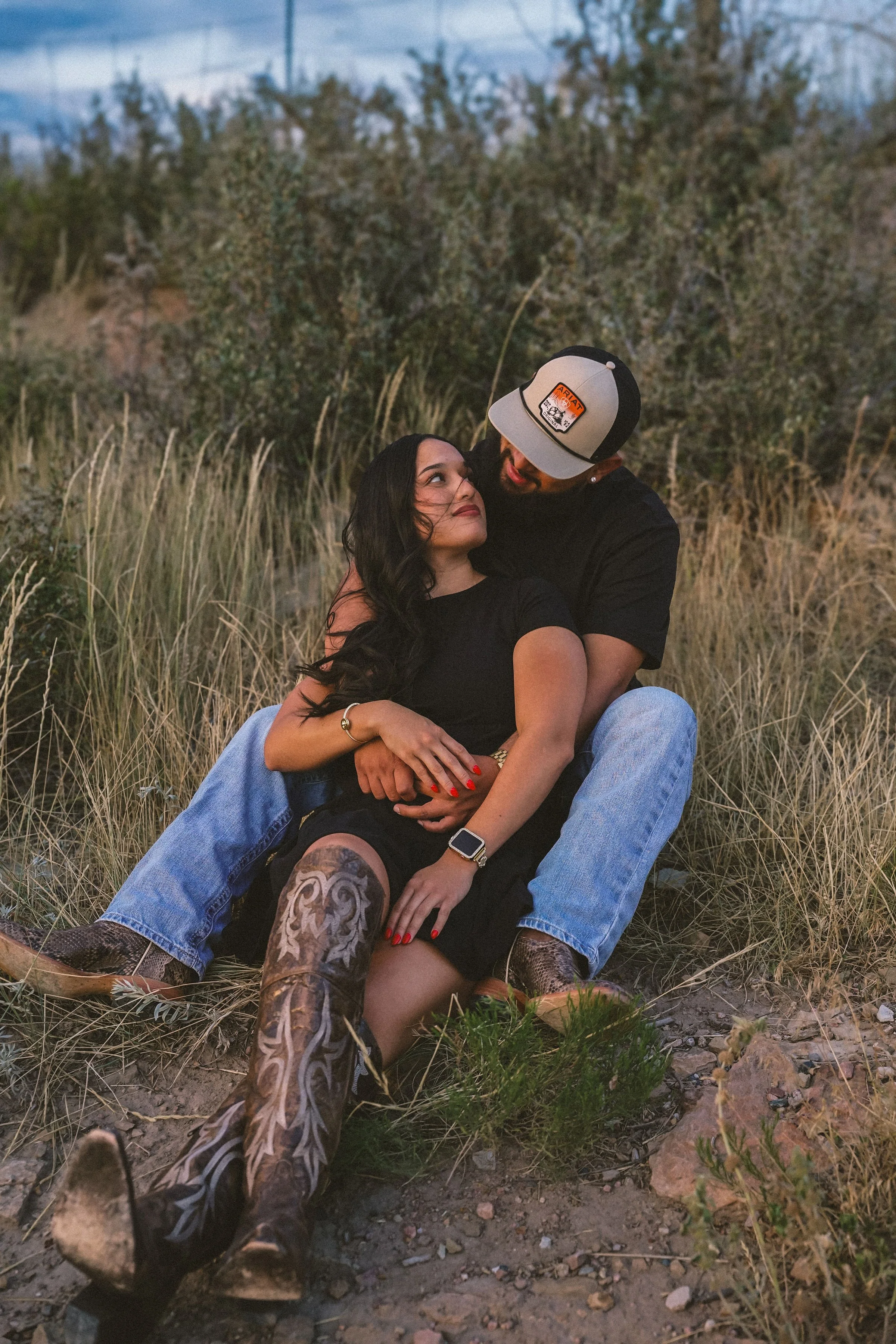 A couple sitting outdoors on the ground surrounded by grass and bushes, with the man embracing the woman from behind as they look into each other's eyes, near sunset or dusk.