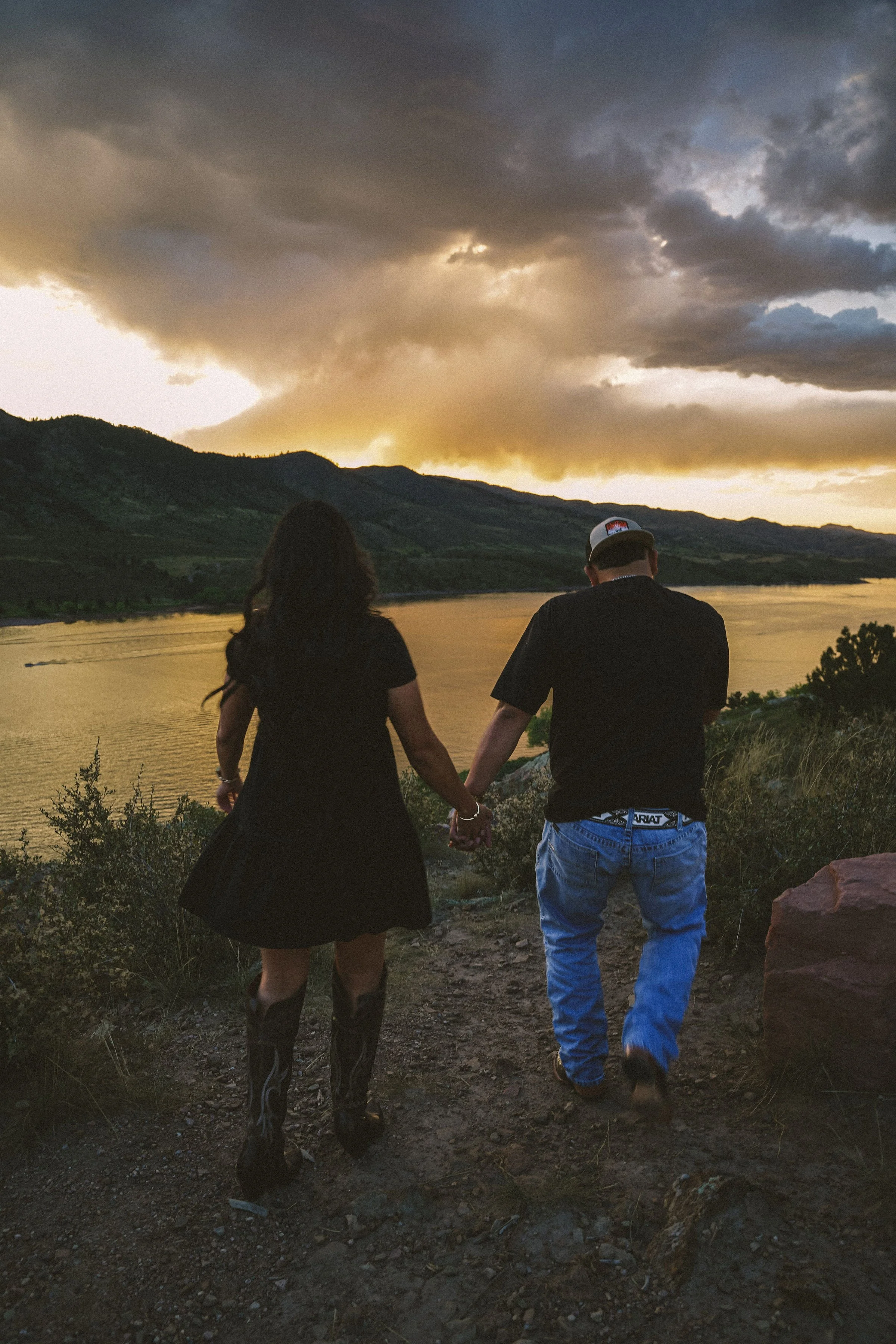 A couple holding hands, standing on a trail overlooking a river and mountains during sunset, with dark clouds overhead.