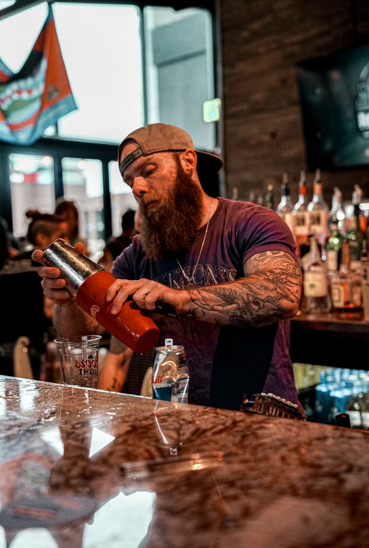A bearded bartender in a backward baseball cap, wearing a dark t-shirt, pours a drink into a glass behind a bar with a granite countertop. Behind him are various liquor bottles on shelves, and there are people in the background inside a bar or restau