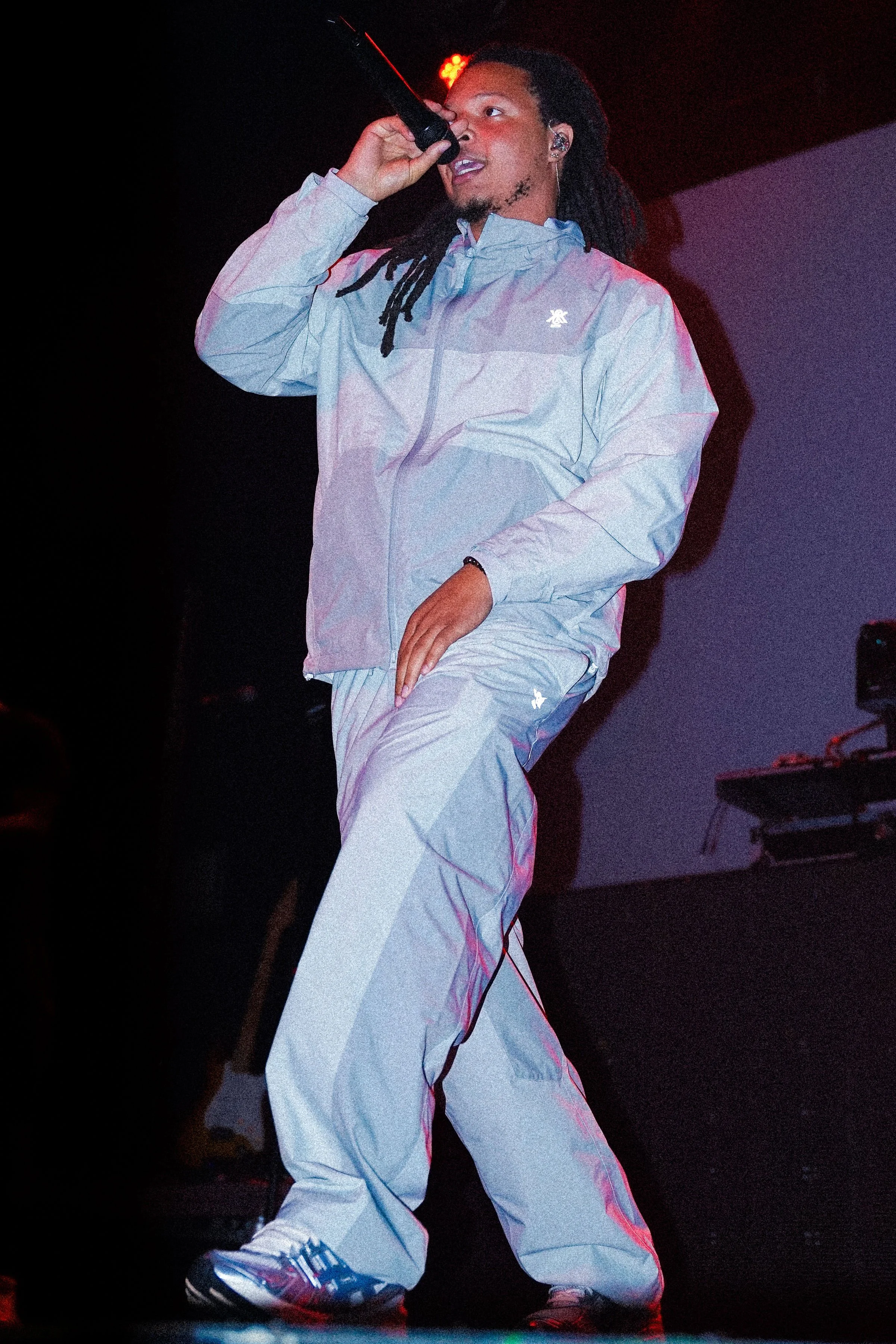 A man with dreadlocks singing into a microphone on stage, wearing a light-colored athletic jacket and matching pants, with a dark background and stage lighting.