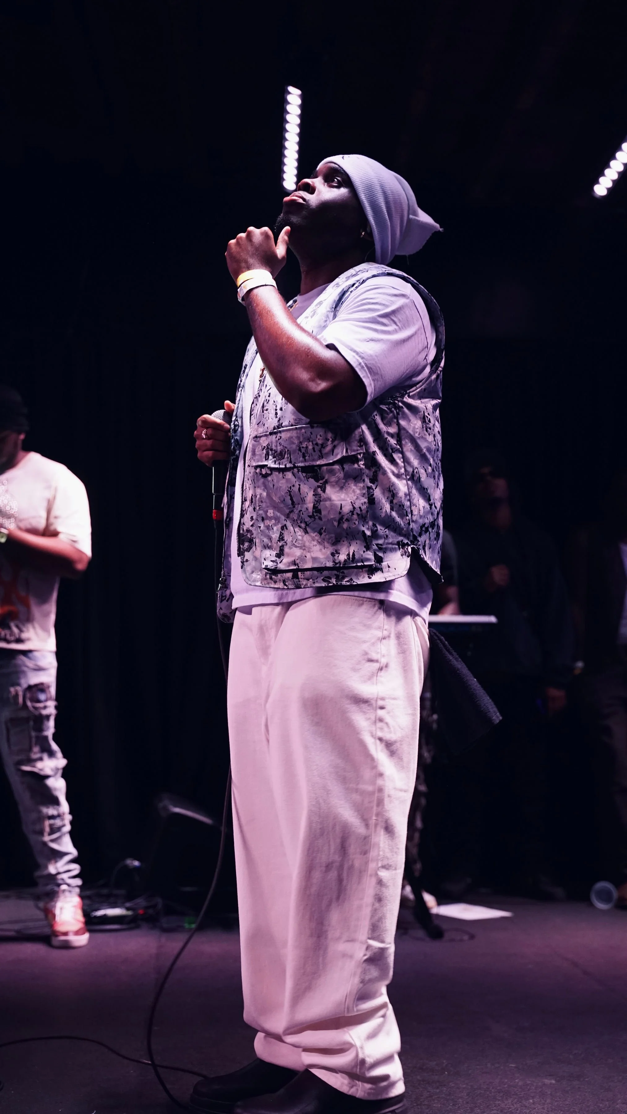 A man standing on stage, wearing a gray beanie, a patterned vest, and white pants, thinking or observing. There are other people in the background and stage lighting.