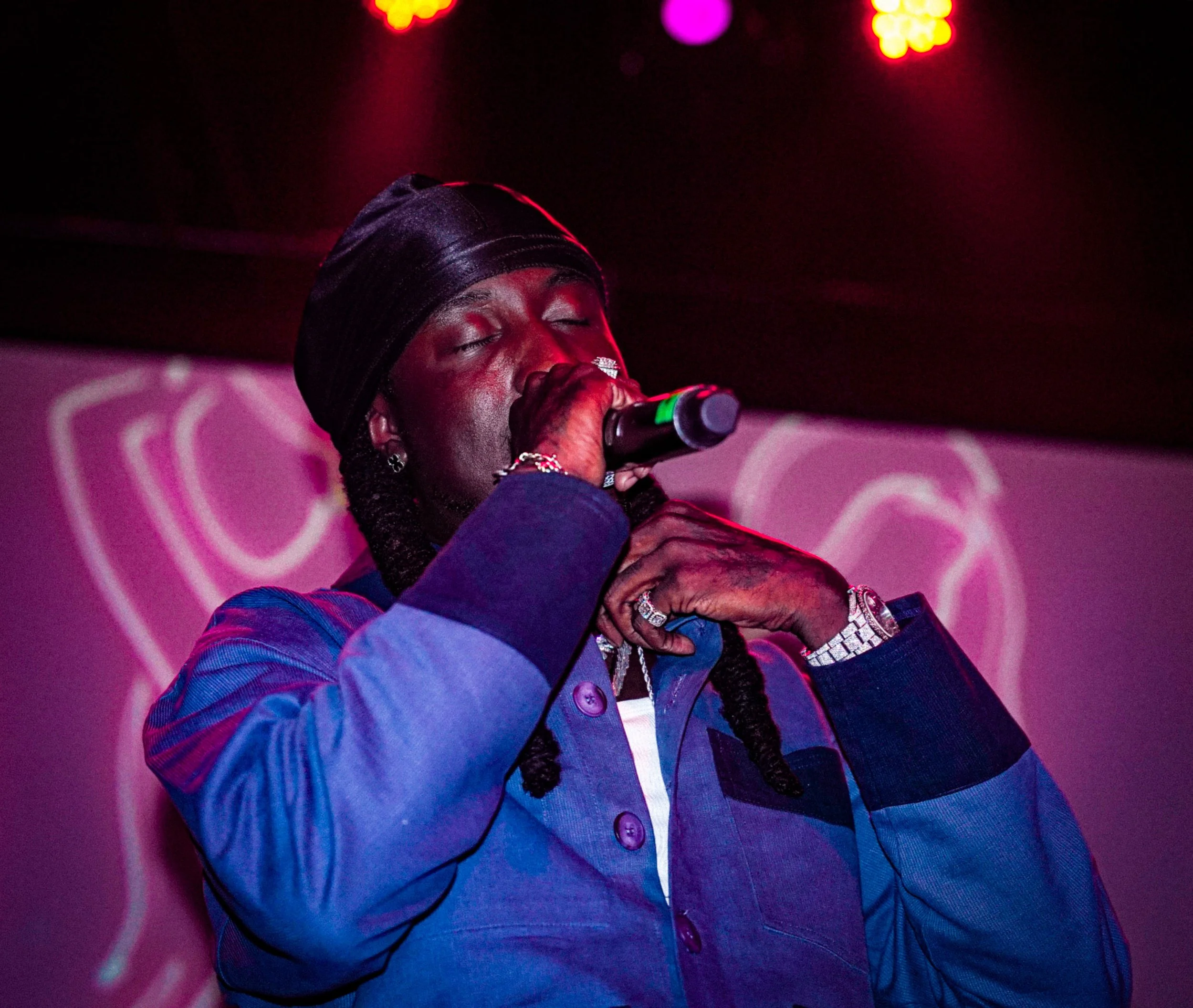 A person with dark skin wearing a black hat and a colorful jacket is singing into a microphone on stage. The background has pink and purple lighting with a heart-shaped graphic.
