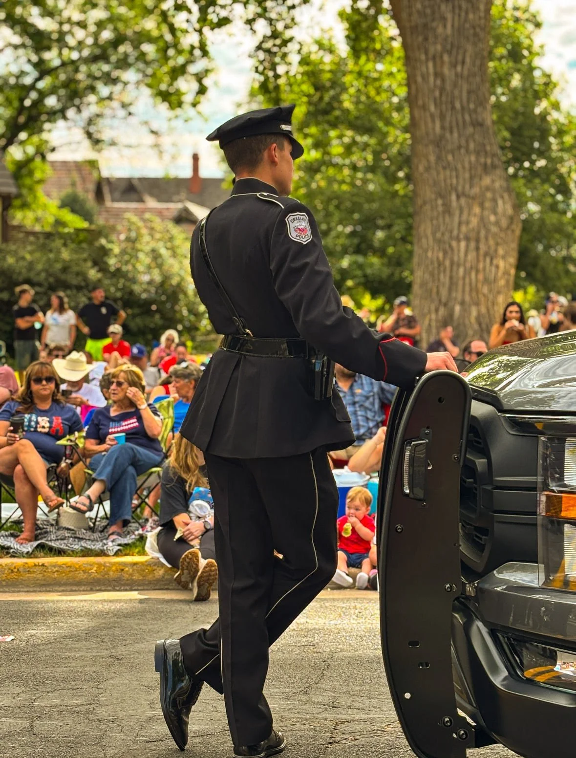 A police officer in uniform stands in front of a black police vehicle at an outdoor event, with a crowd of seated and standing people under trees in the background.
