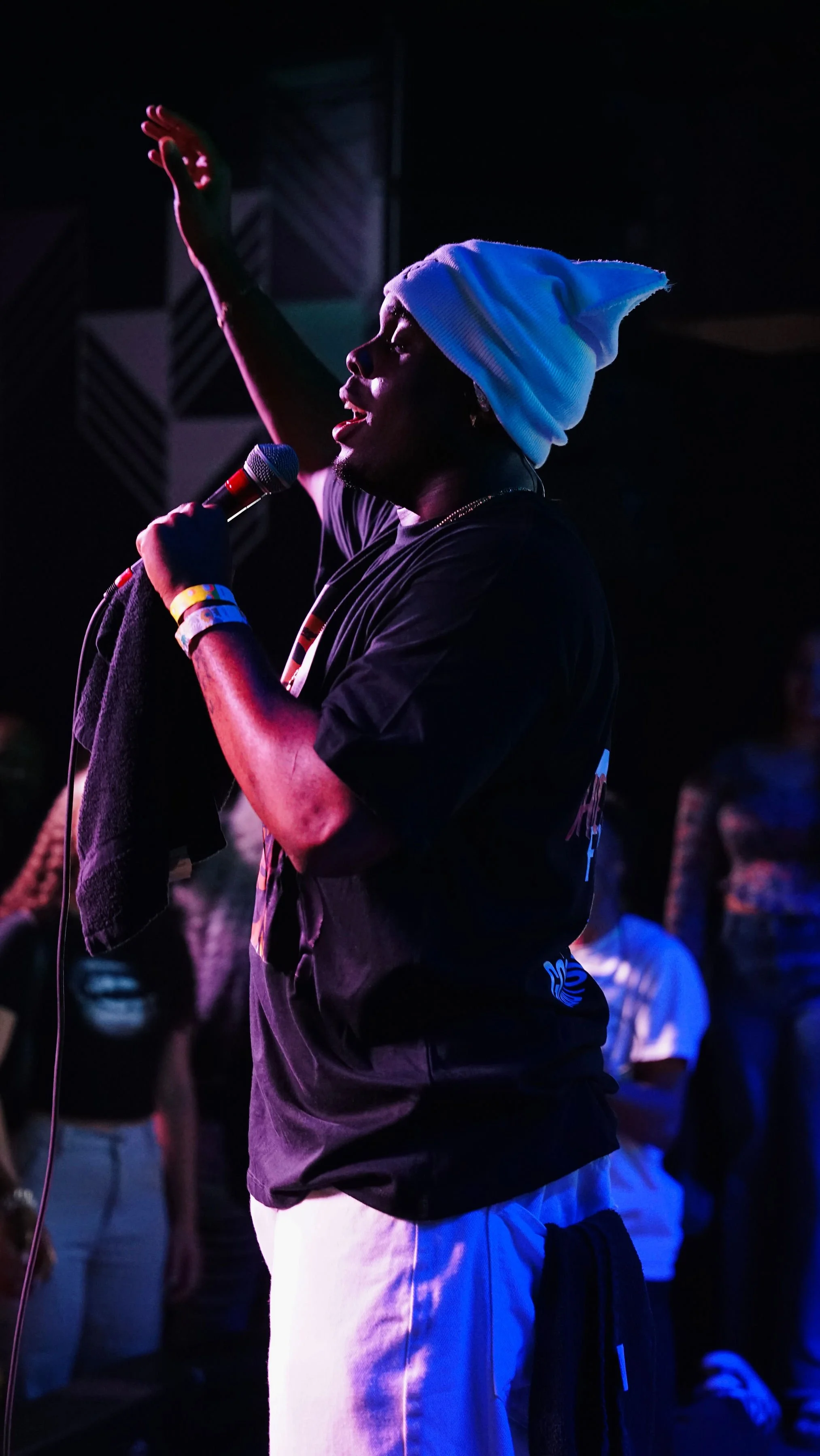 A person wearing a white beanie and dark T-shirt, holding a microphone and singing, with their arm raised, in a dimly lit setting with a crowd in the background.
