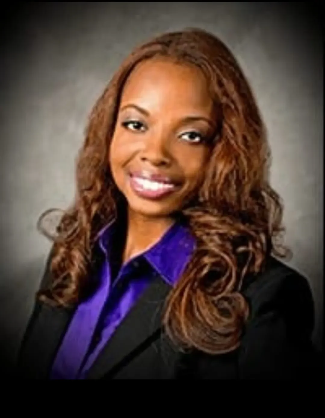 A professional woman with long, wavy, reddish-brown hair, wearing a black blazer and a purple blouse, smiling at the camera against a plain gray background.