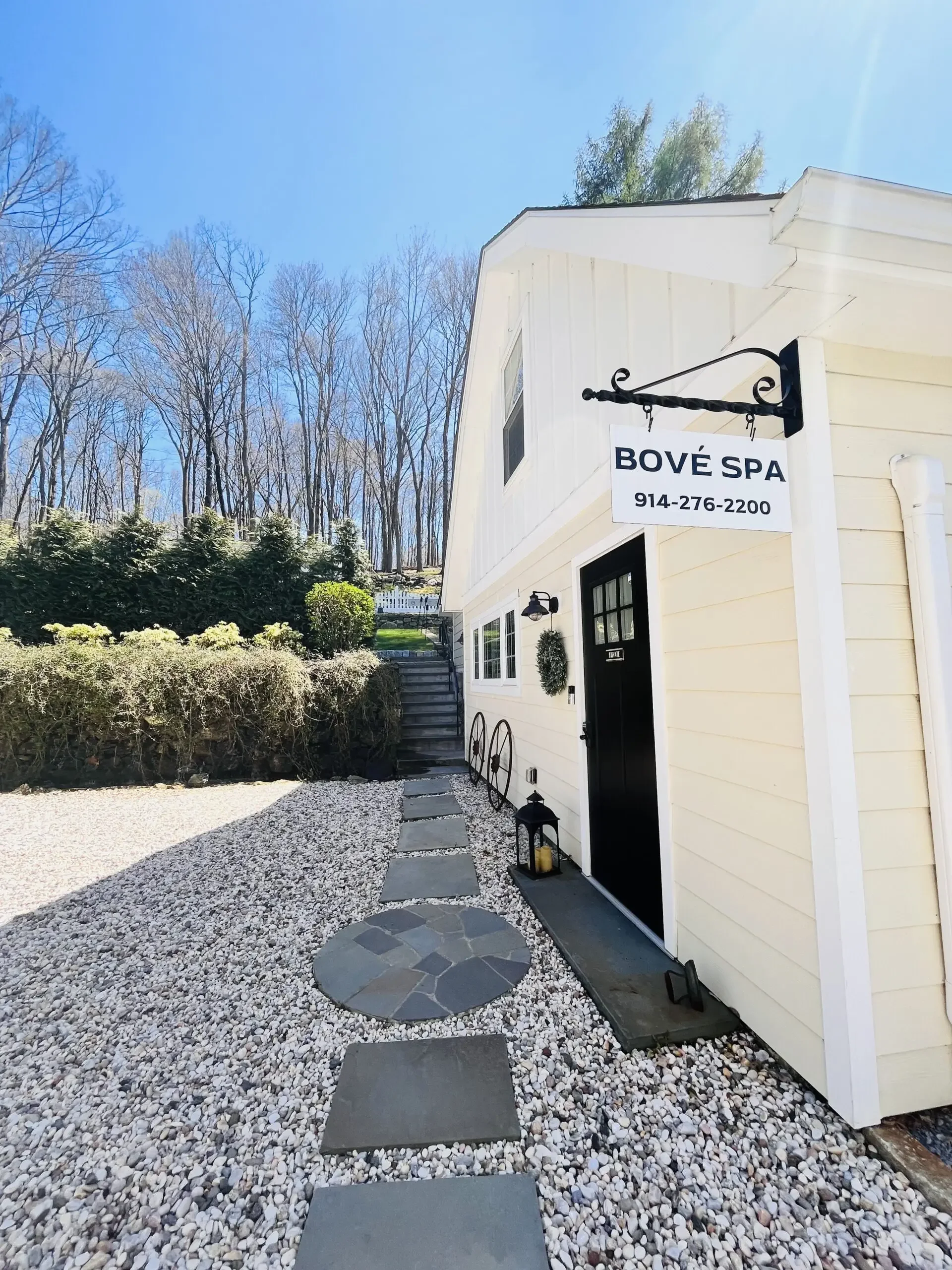 White building with a sign reading 'BOVÉ SPA' and a phone number, black door, outdoor lanterns, and stone pathway with gravel leading upward, surrounded by leafless trees and bushes on a sunny day.