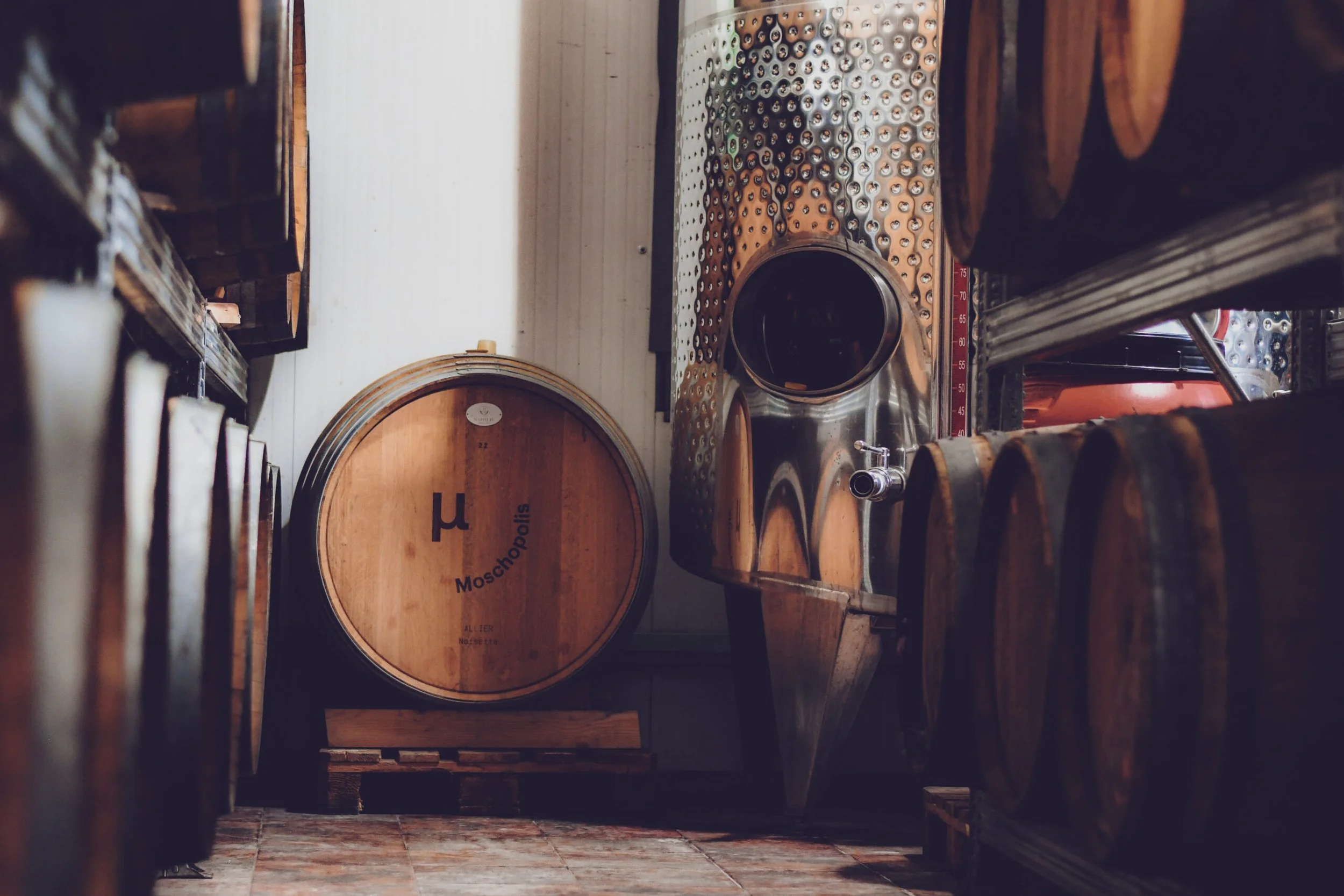 View of wine barrels stored in the Moschopolis cellar with one visible metal tank on the right side, and a wooden floor.