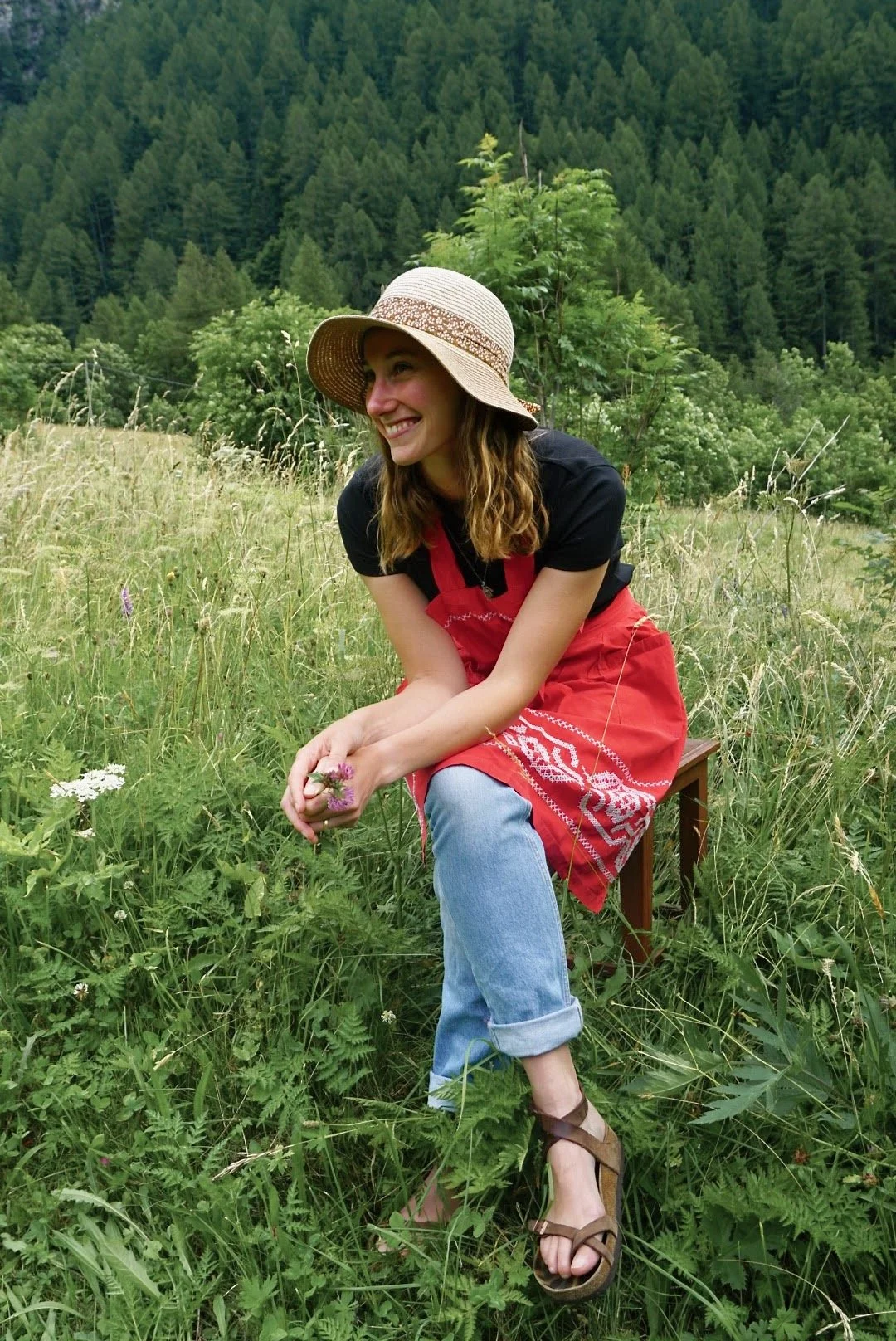 Sarah sitzt mit Sonnenhut und Schürze auf einer Bank auf einer Wiese