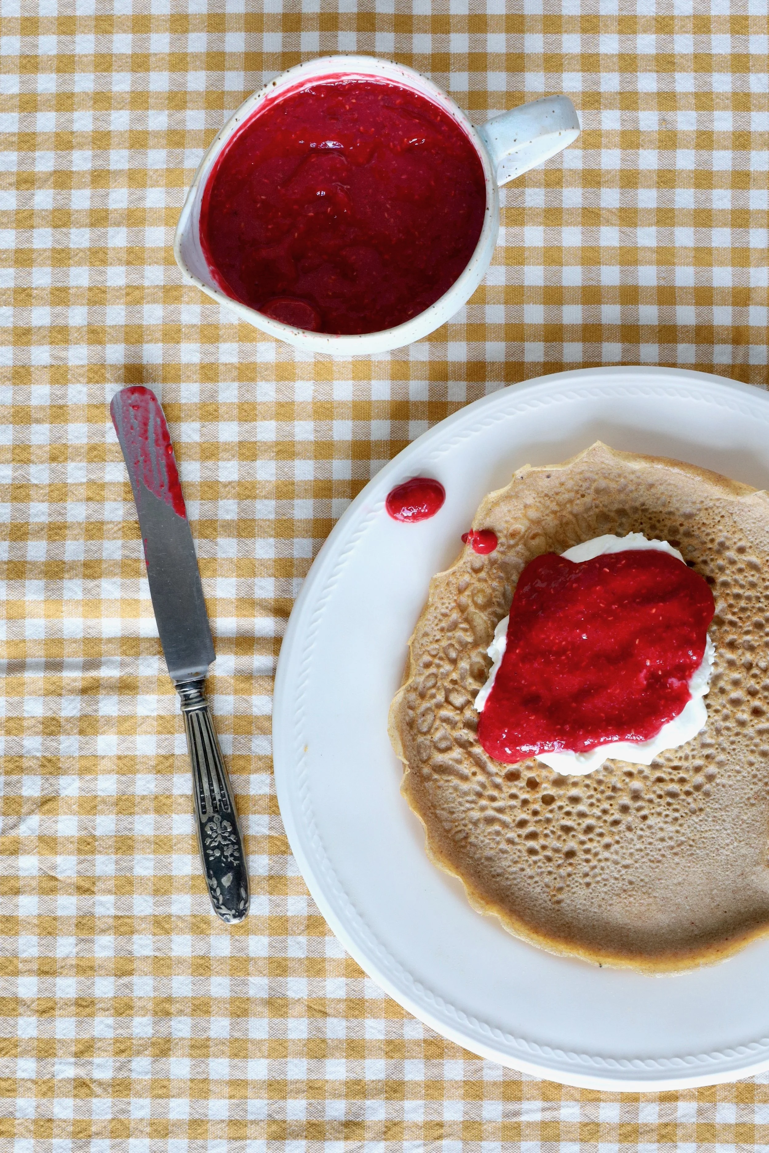 Ein Glas Himbeermarmelade neben einem Teller mit einem Pfannkuchen, auf dem Himbeermarmelade liegt