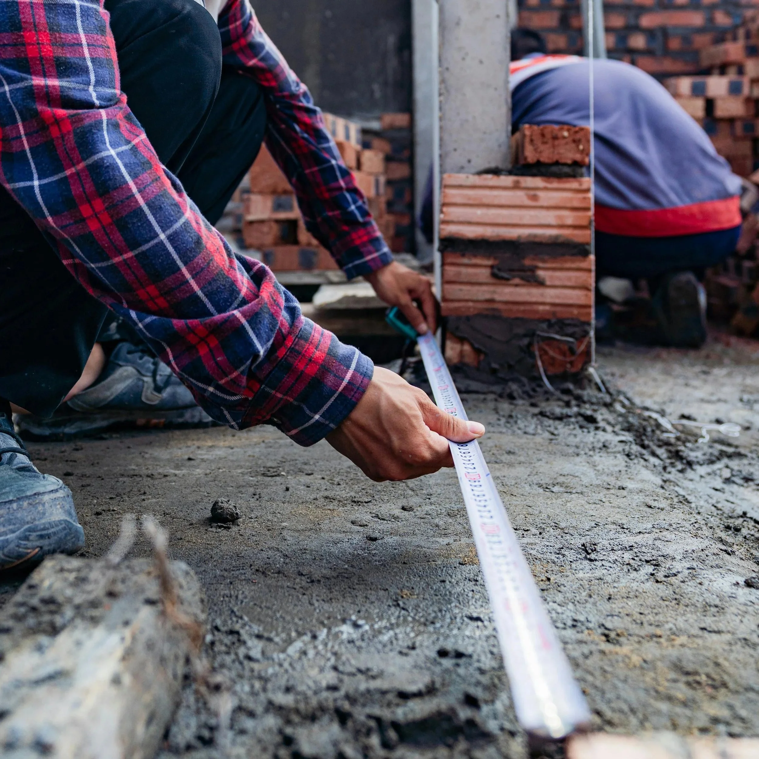 Two construction workers measuring the ground at a construction site with a tape measure. bricks and construction materials. free quote, onsite consultation, layout, pricing, options