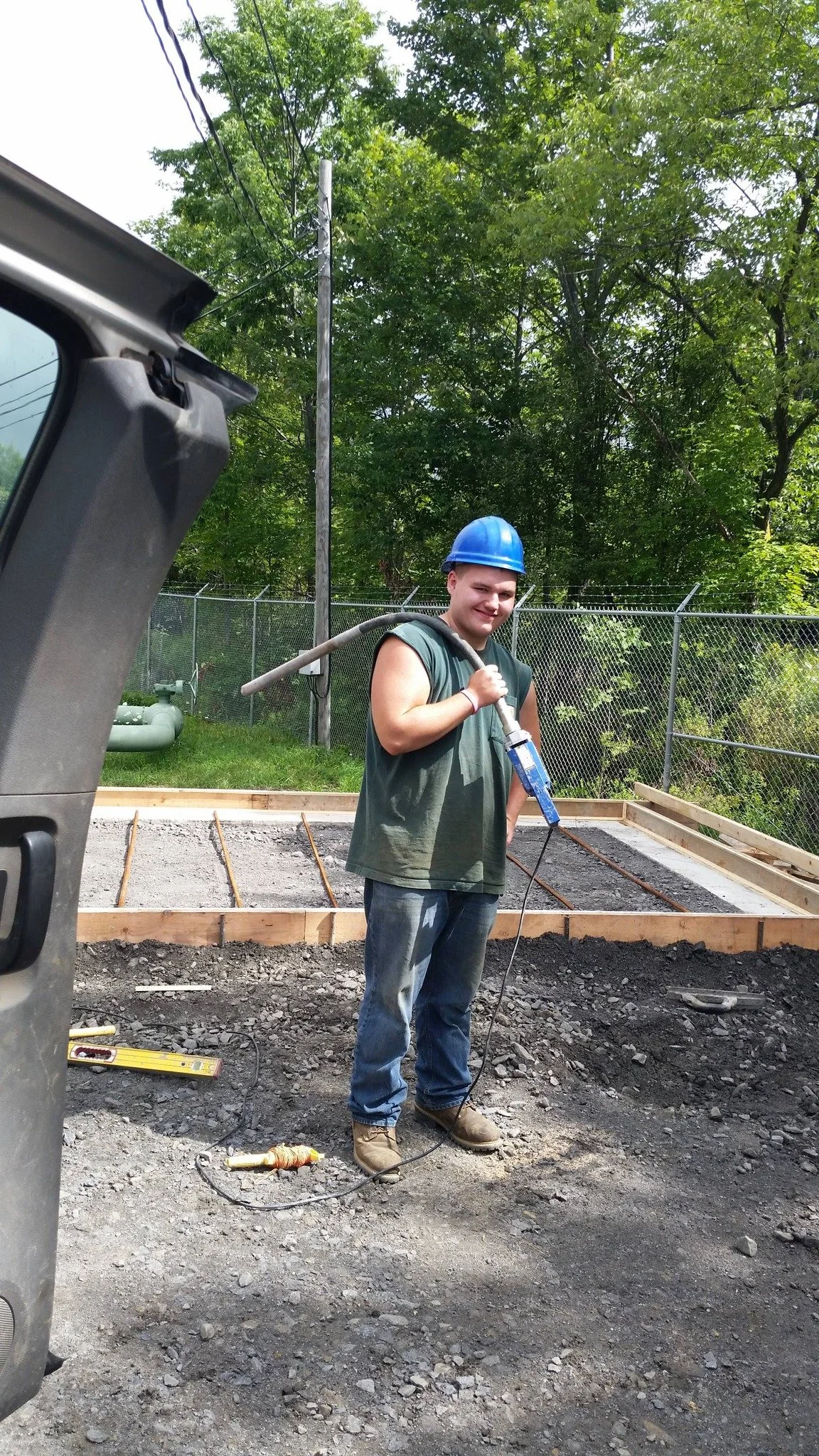 A young man wearing a blue construction helmet and a sleeveless shirt is working on a construction site, holding a power tool. There are wooden frames and construction supplies on the ground, with a green, leafy background and a chain-link fence.