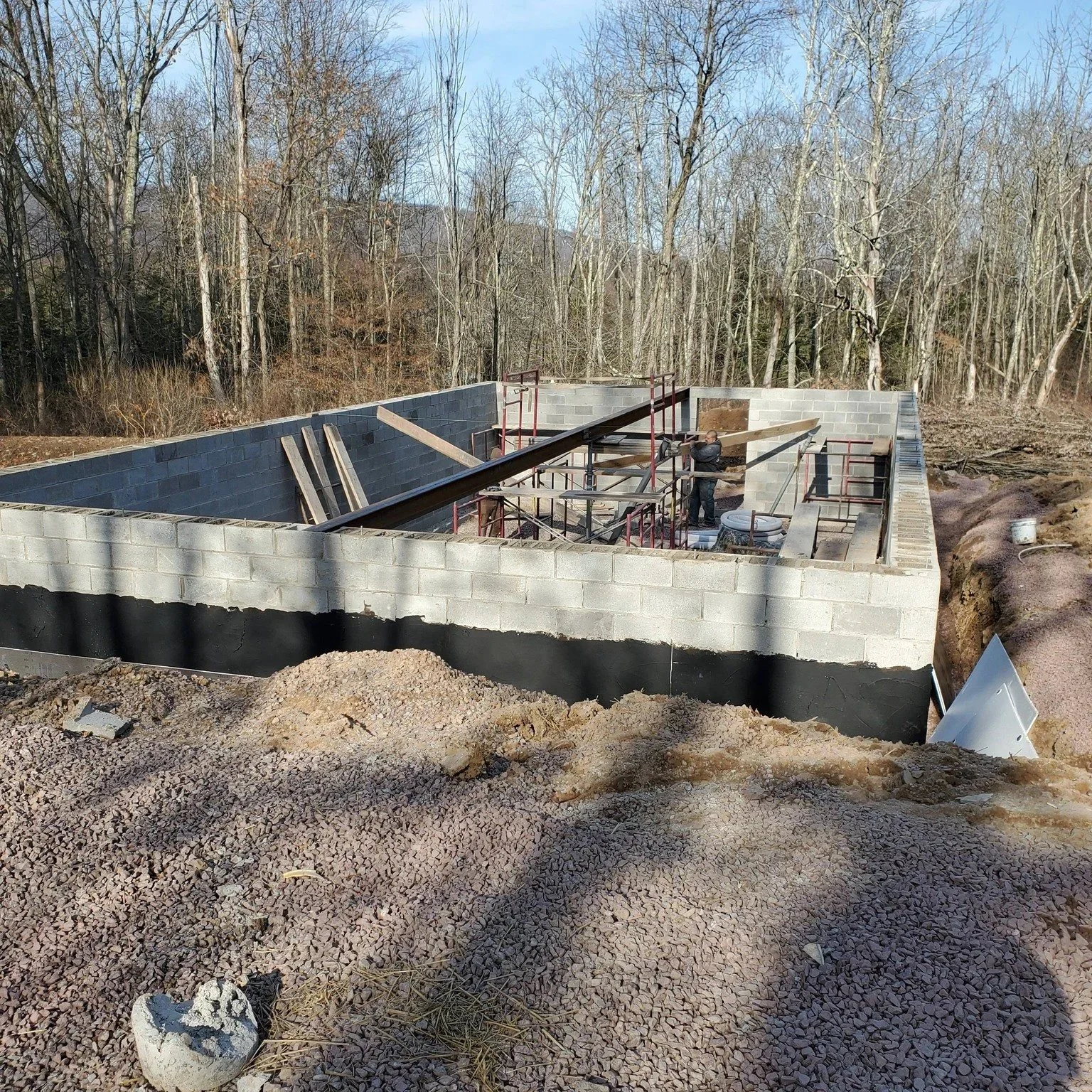 Construction site with concrete block foundation and steel beams, surrounded by trees, and a worker installing structural elements. water proofing, drainage. excavation, footers, posts, grade