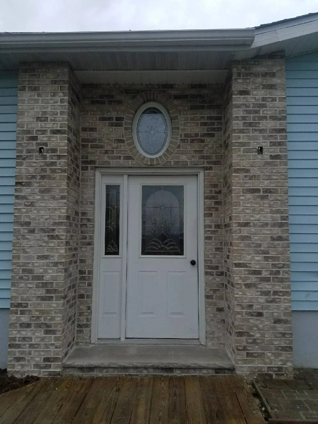 Front door of a house with brick trim and a stained glass window, with a wooden porch in front. stone trim, facade, wainscot