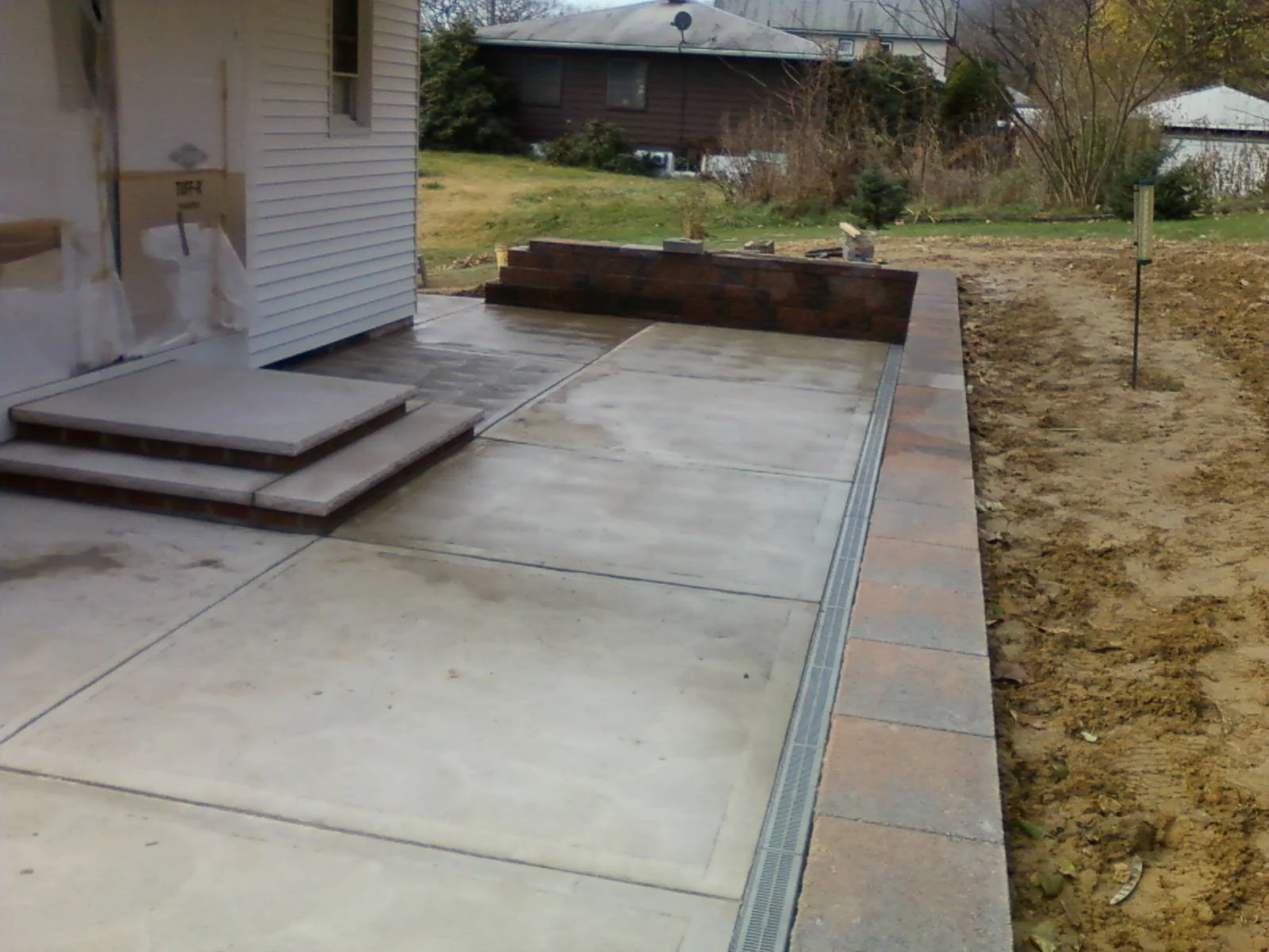 A newly paved concrete patio with a brick border next to a house, with four small steps leading to the house doorway and a retaining wall at the back of the patio, along with some construction tools and materials nearby.