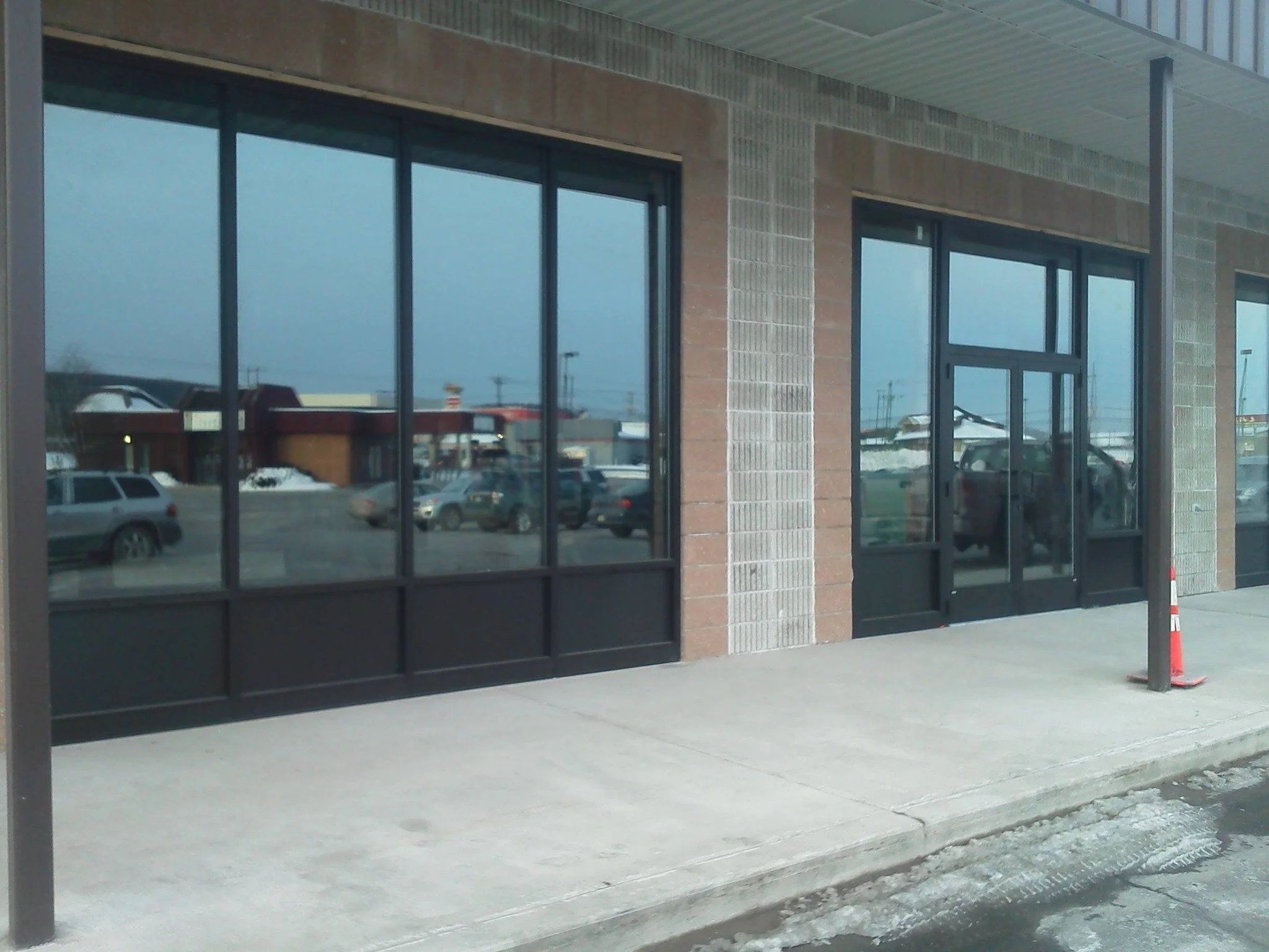 Empty storefront with large glass windows and a glass door, parking lot with cars nearby, snowy ground, and a construction cone outside.