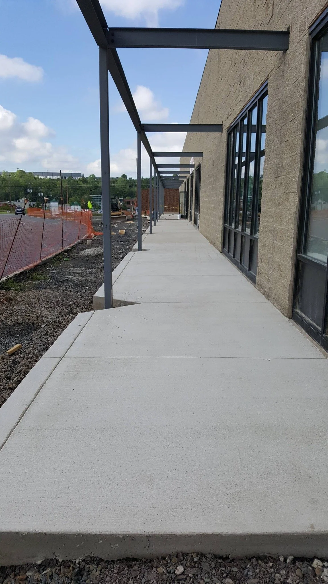 Newly constructed concrete walkway outside a commercial building with large window panels, covered partly by metal awnings, with ongoing construction visible in the background.