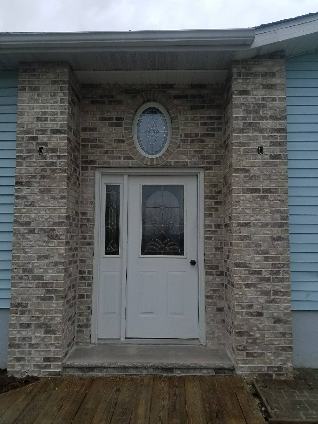 Front entrance of a house with a white door and window, brick and blue siding exterior, and a decorative oval window above the door.