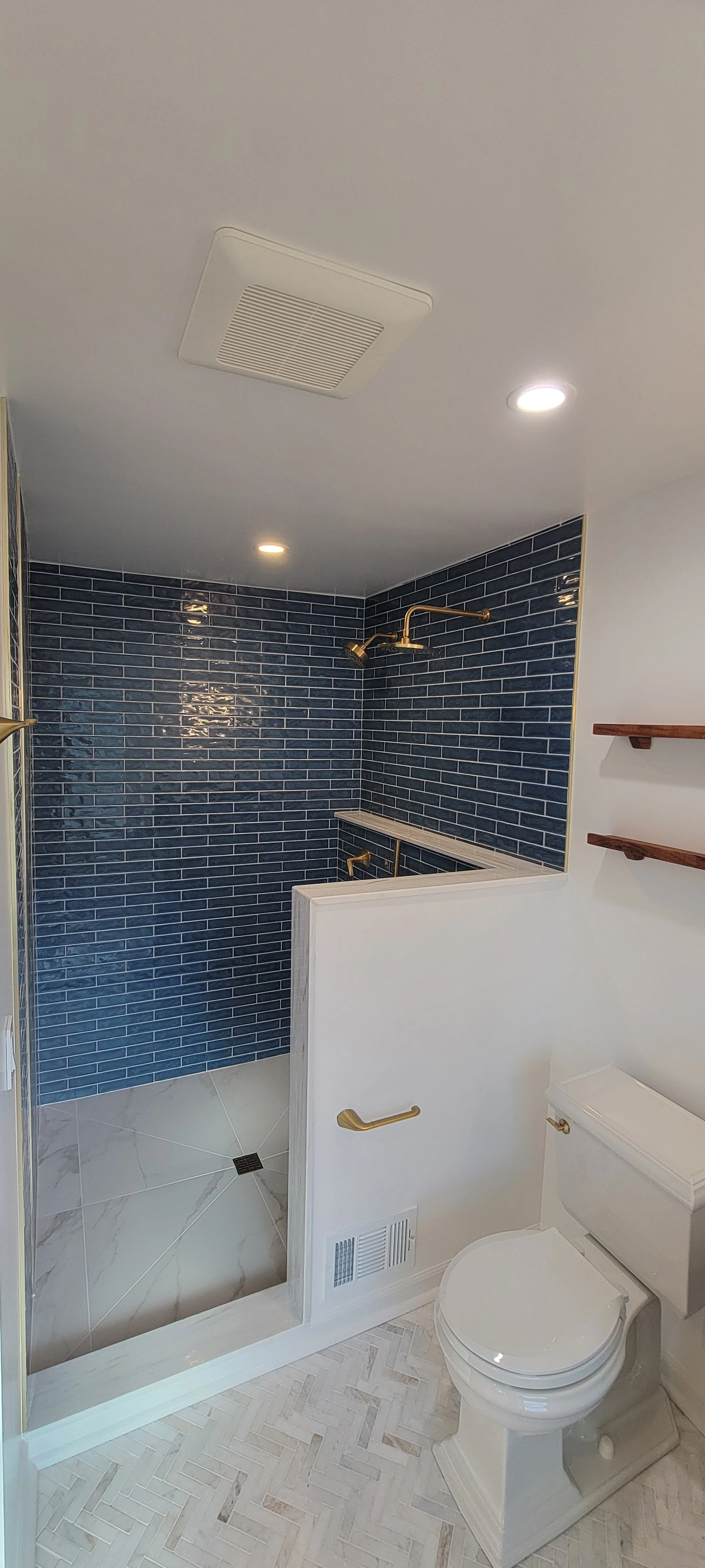 Bathroom with a walk-in shower featuring dark blue subway tiles, a brass showerhead, and a partial white wall with brass grab bars, next to a white toilet and wooden shelves.