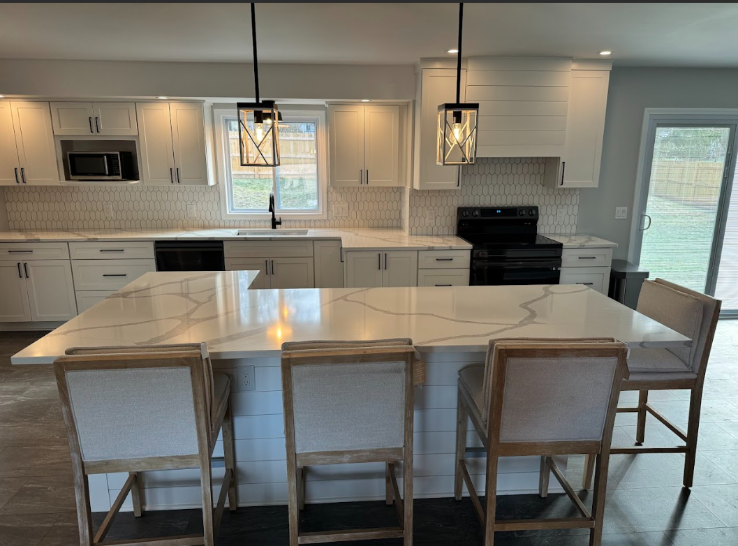 Modern kitchen with white cabinets, a marble countertop island with seating, black appliances, pendant lights, and a sliding glass door leading outside.