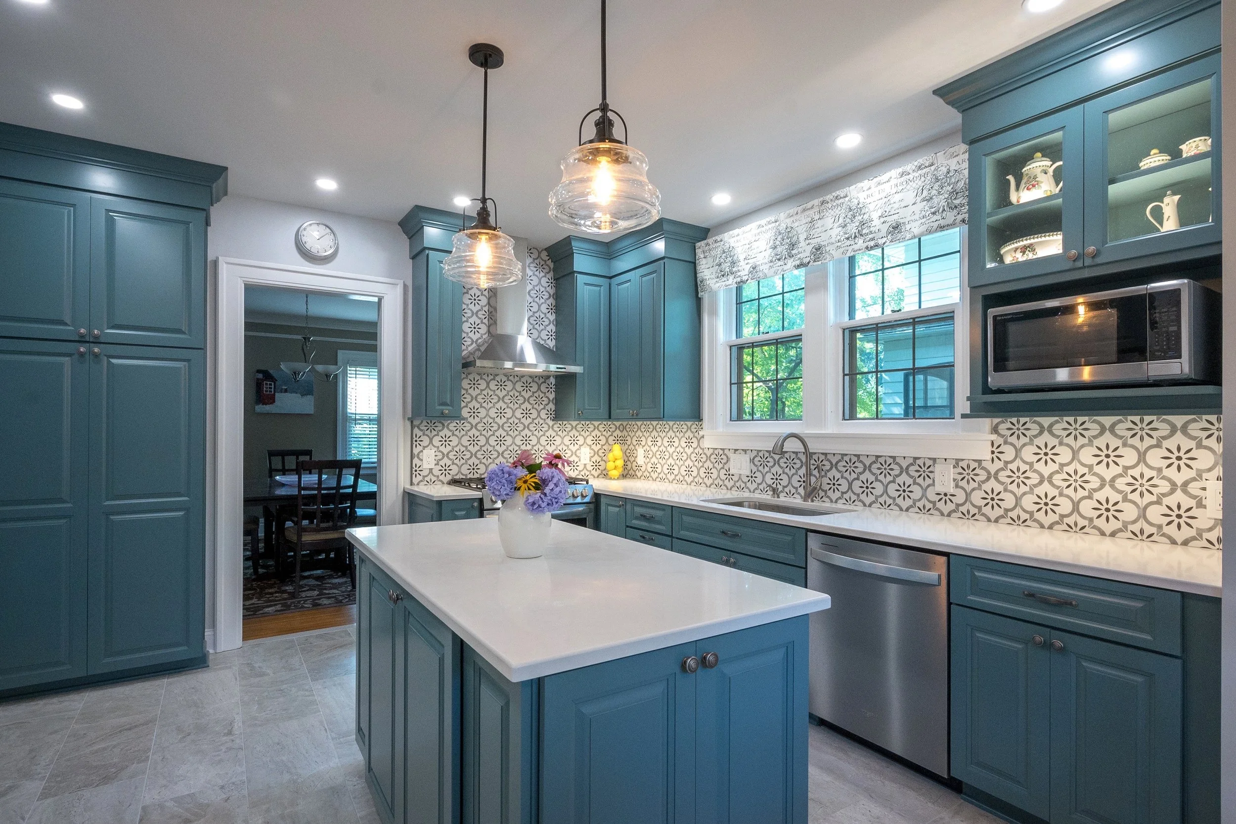 Kitchen with blue cabinets, white countertop island, patterned tile backsplash, stainless steel appliances, and a vase with purple, yellow, and pink flowers on the island.