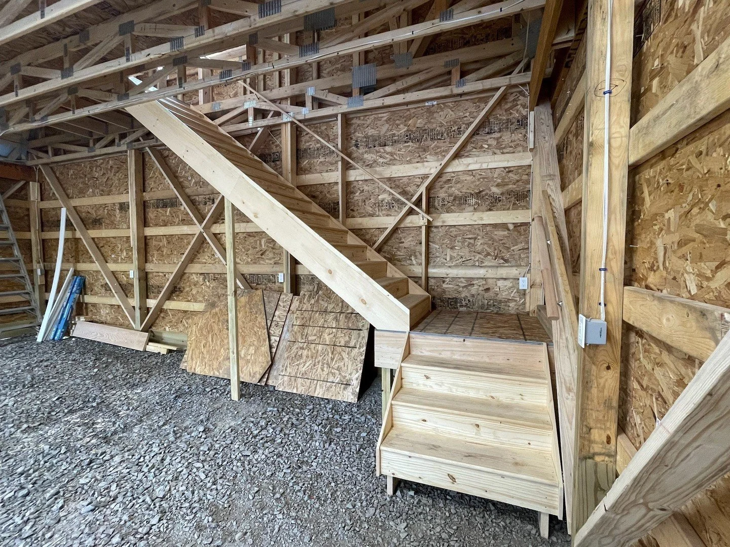 Wooden staircase under construction inside a building, with exposed wood framing and gravel floor.