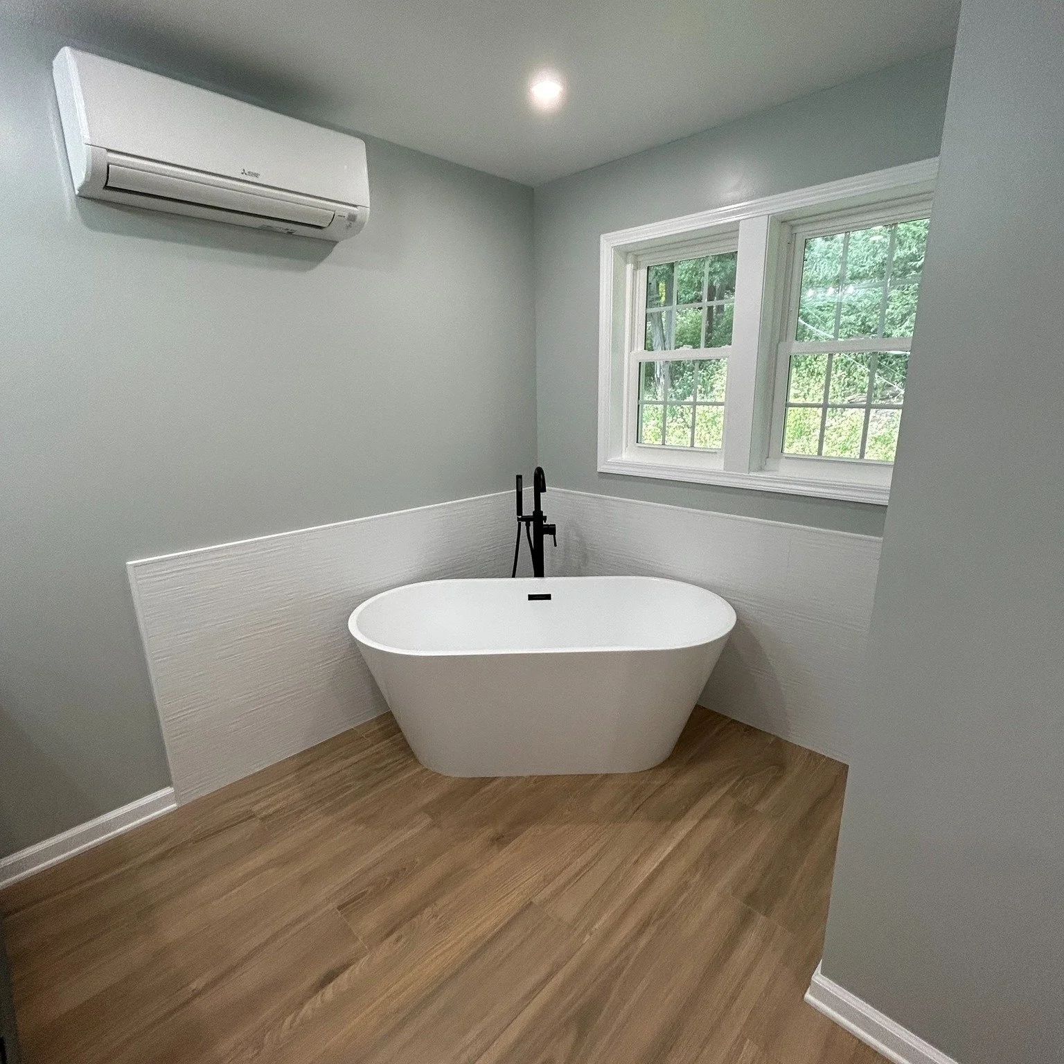 Modern bathroom with freestanding white bathtub, black floor-mounted faucet, light gray walls, wood-look flooring, a large window, and a wall-mounted air conditioner.