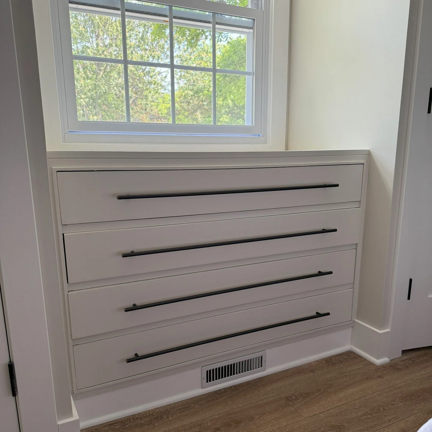 White dresser with five drawers and black handles, situated beneath a window showing green trees outside, located in a room with white walls and hardwood flooring.