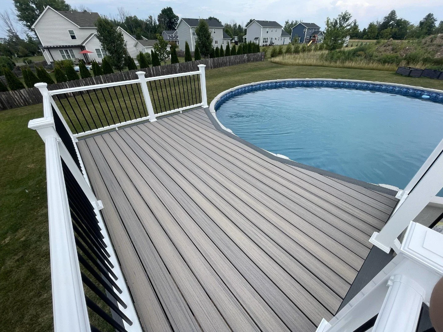 View of a backyard with a above-ground swimming pool, a wooden deck, and a wooden fence. Several modern houses are in the background, and the yard is surrounded by grass and trees.