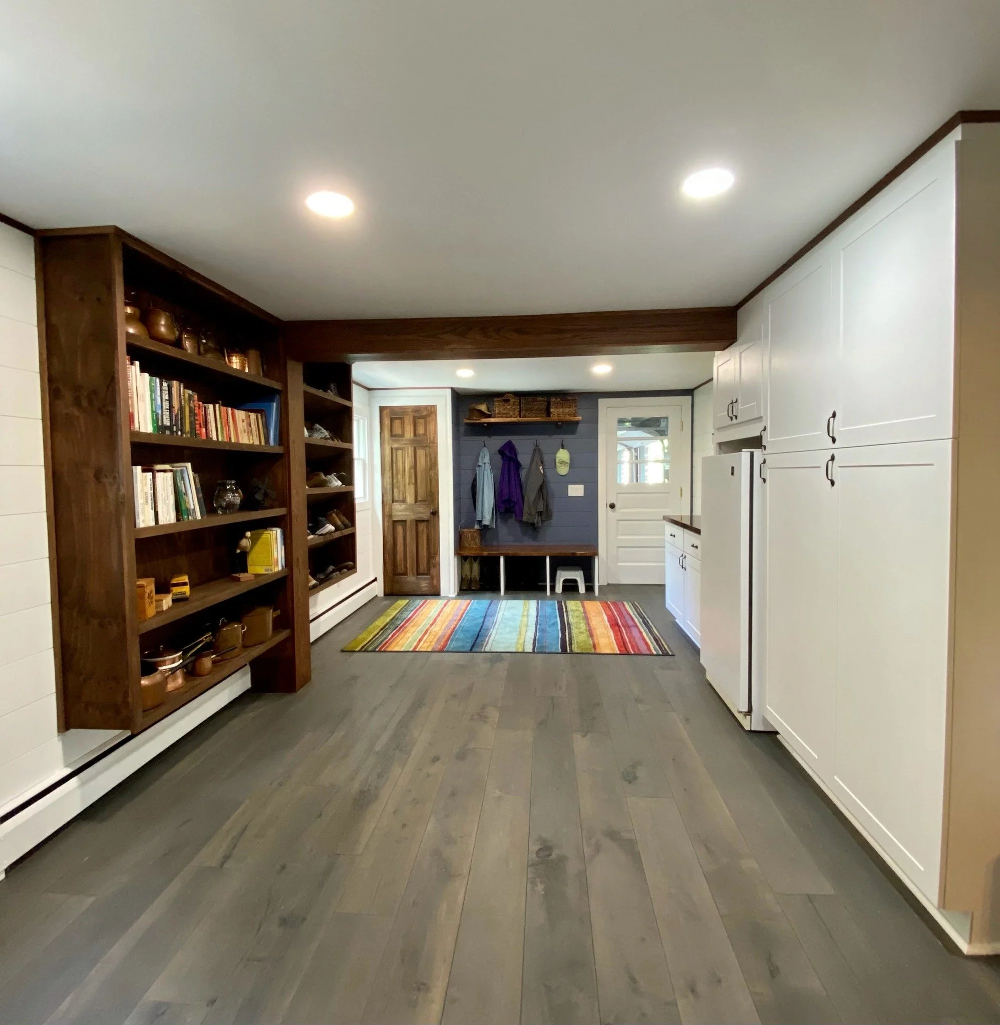 Interior view of a cozy mudroom with white cabinetry, a multicolored striped rug, wooden flooring, and a blue accent wall with hooks and a bench for shoes and coats.