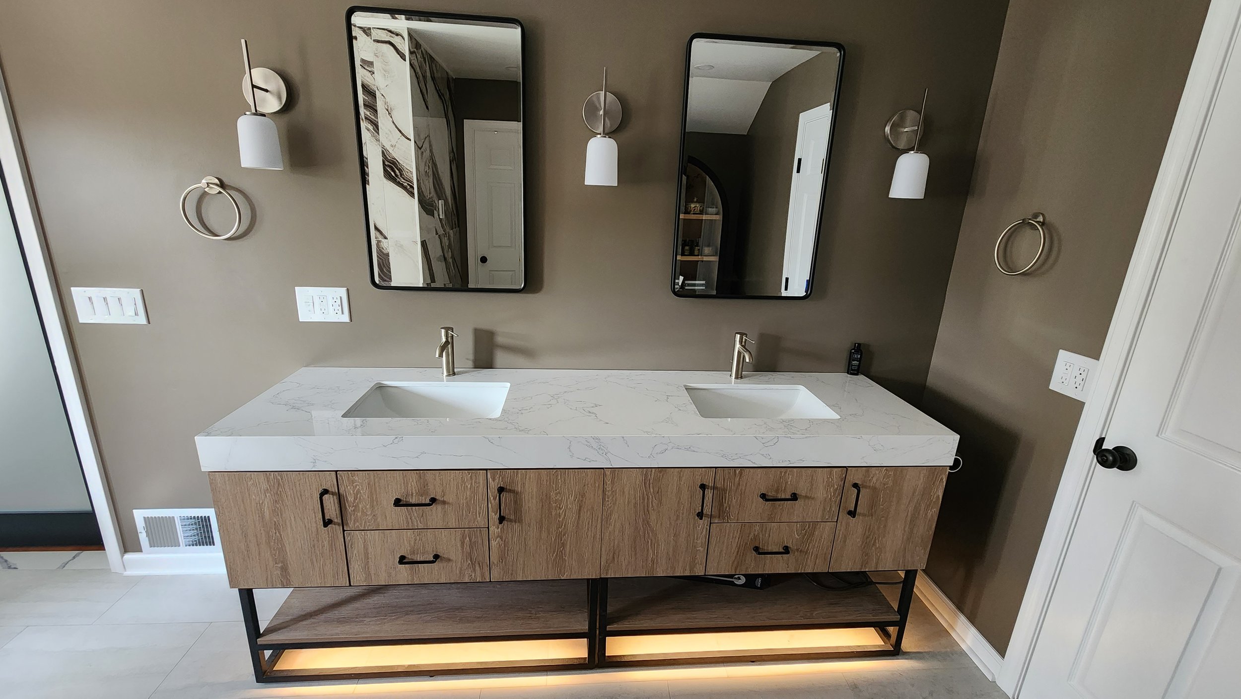 Double bathroom vanity with white marble countertop, two undermount sinks, and wooden cabinet with black handles. Two rectangular mirrors above each sink, with wall-mounted white sconces on either side. Brown wall with multiple electrical outlets and a towel ring on each side.