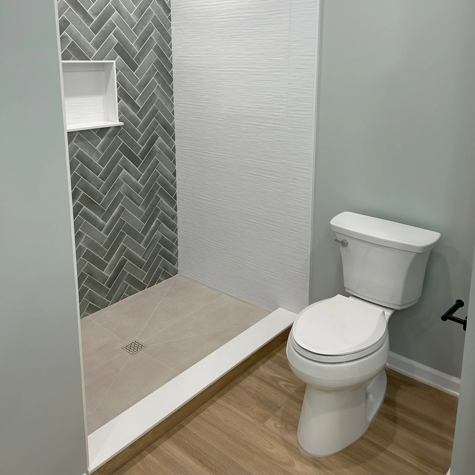 Bathroom with toilet and walk-in shower featuring gray herringbone tile accent wall, white built-in shelf, and wood-look flooring.