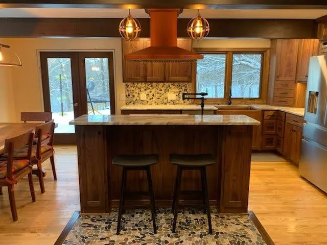 A cozy kitchen with wooden cabinets, a kitchen island with two stools, a granite countertop, a backsplash with a floral pattern, a double oven, and large windows showing trees outside. There are pendant lights hanging above the island and a sliding g