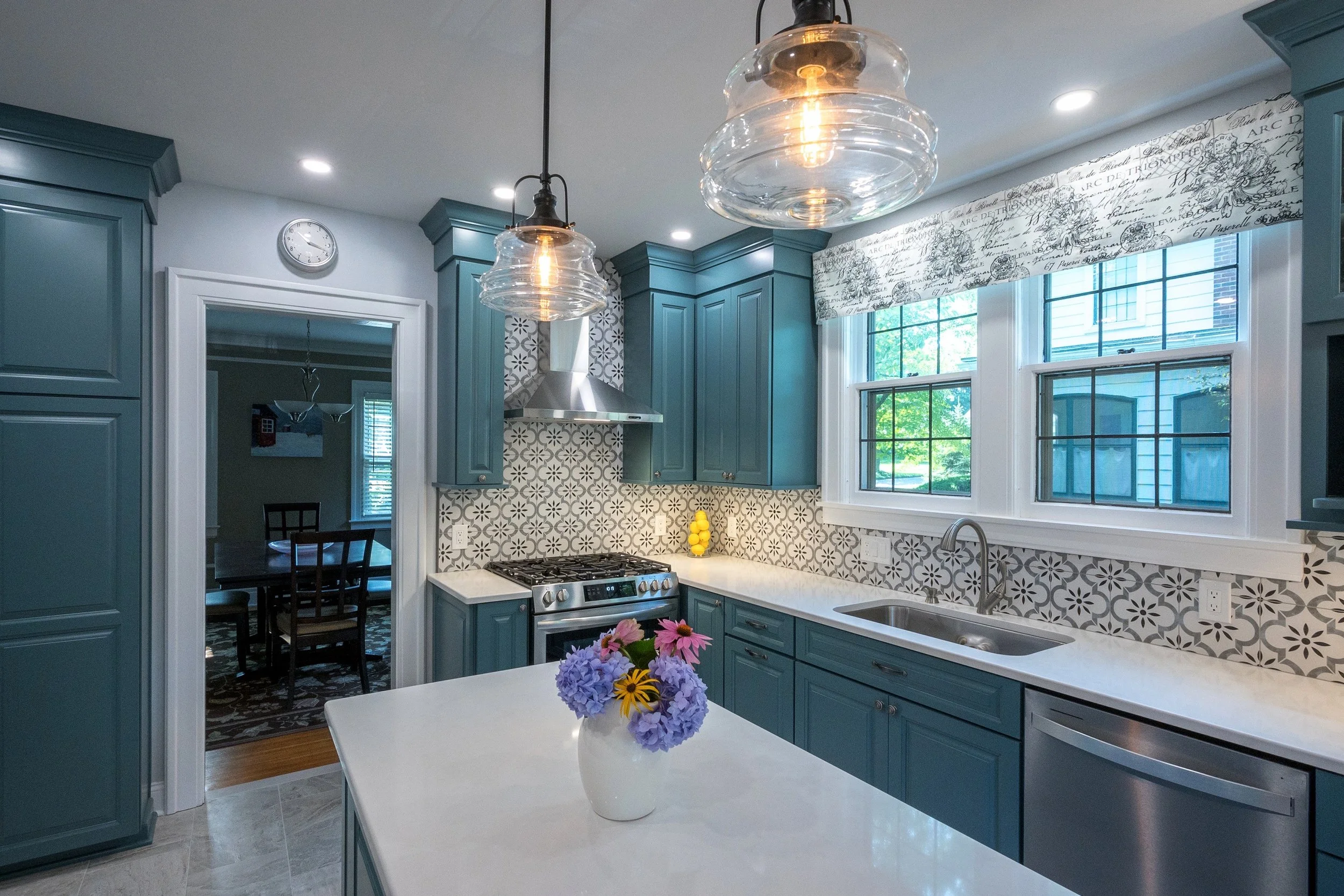 Modern kitchen with blue cabinets, white countertops, patterned backsplash, and a vase of purple, pink, and yellow flowers on the island. Large window, two pendant lights, stainless steel appliances, and a doorway to a dining room.